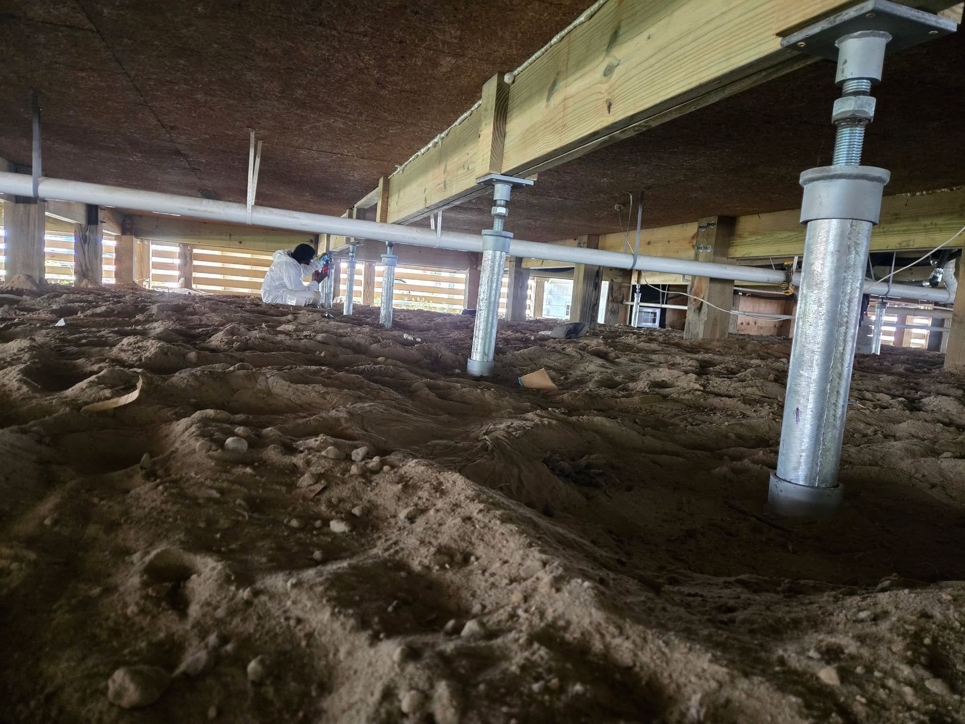 Low crawlspace under a building with dirt floor, white support posts, and beams overhead