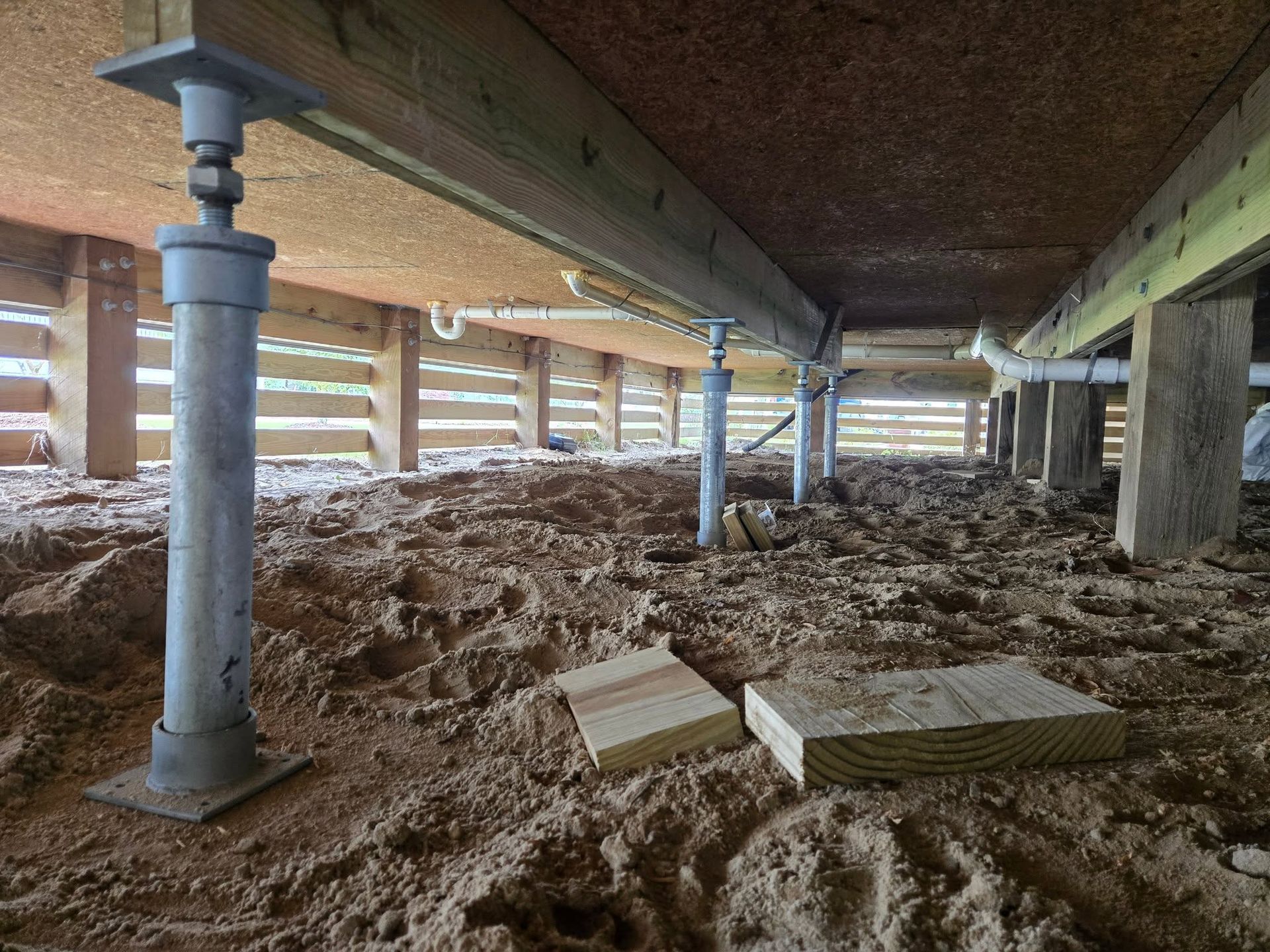 Muddy crawlspace under a building, with support beams, columns, and a small work light.