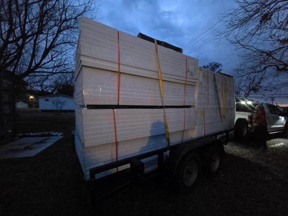Flatbed trailer loaded with stacked lumber, strapped down at dusk in a wooded yard.
