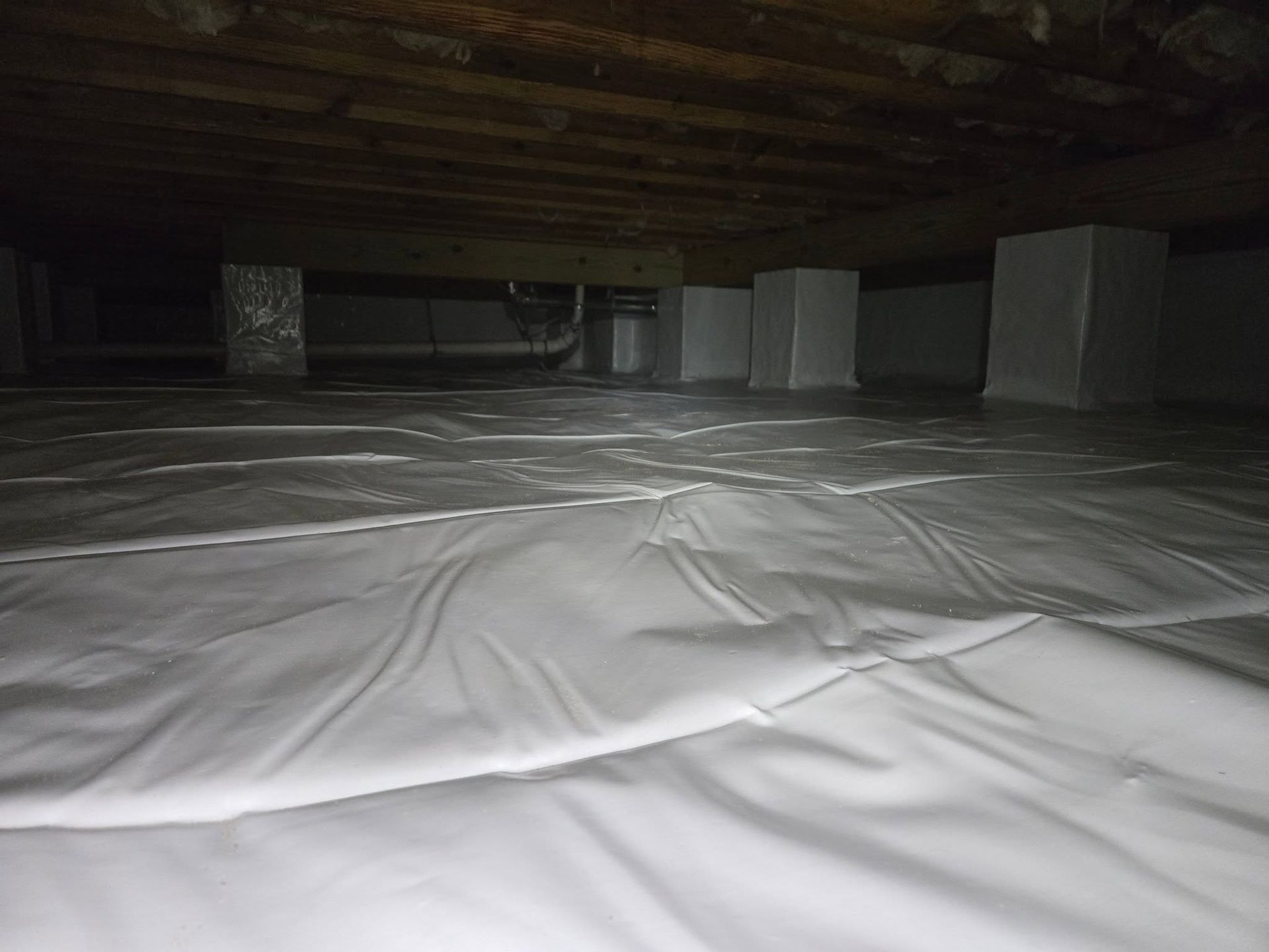 Dim basement with plastic-covered floor and large white support columns under a low wooden ceiling
