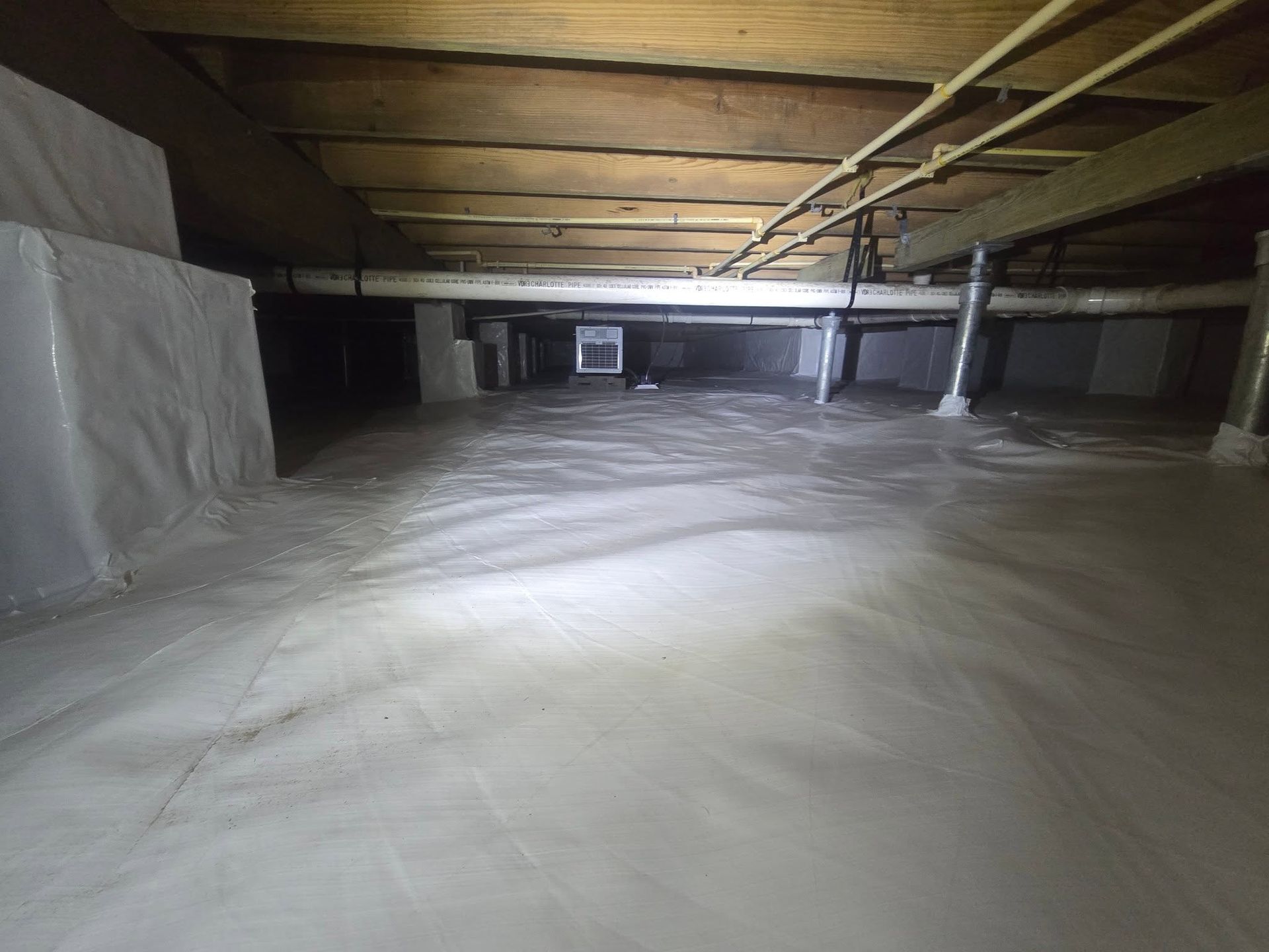 Crawlspace with exposed wooden joists, insulation, and white plastic-covered ground lit by a flashlight
