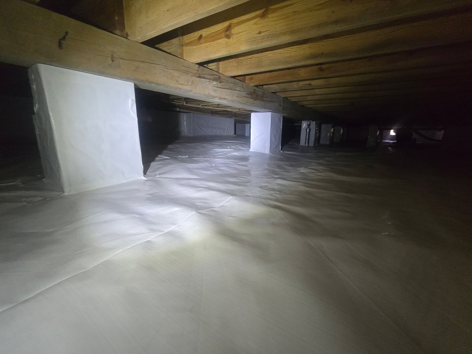 Flooded crawl space under a house with wooden beams and concrete supports, lit by a flashlight