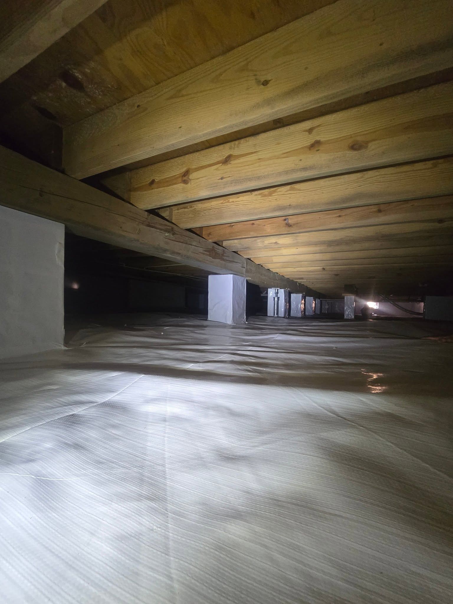 Low wooden crawlspace or attic with exposed beams and a dim, dusty floor lit by a flashlight