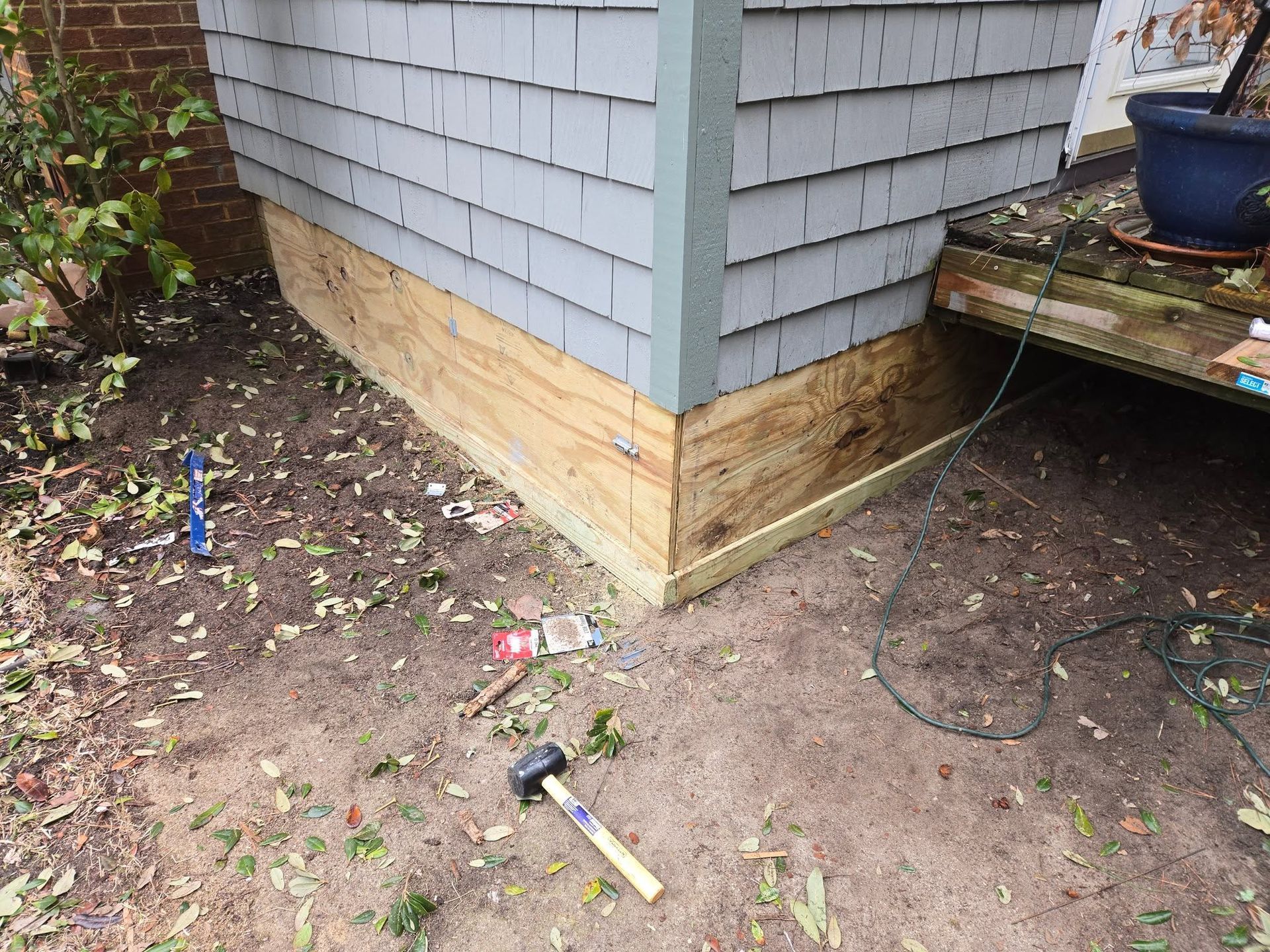 Damage repaired at house corner with new plywood siding; yard tools and debris lie nearby.