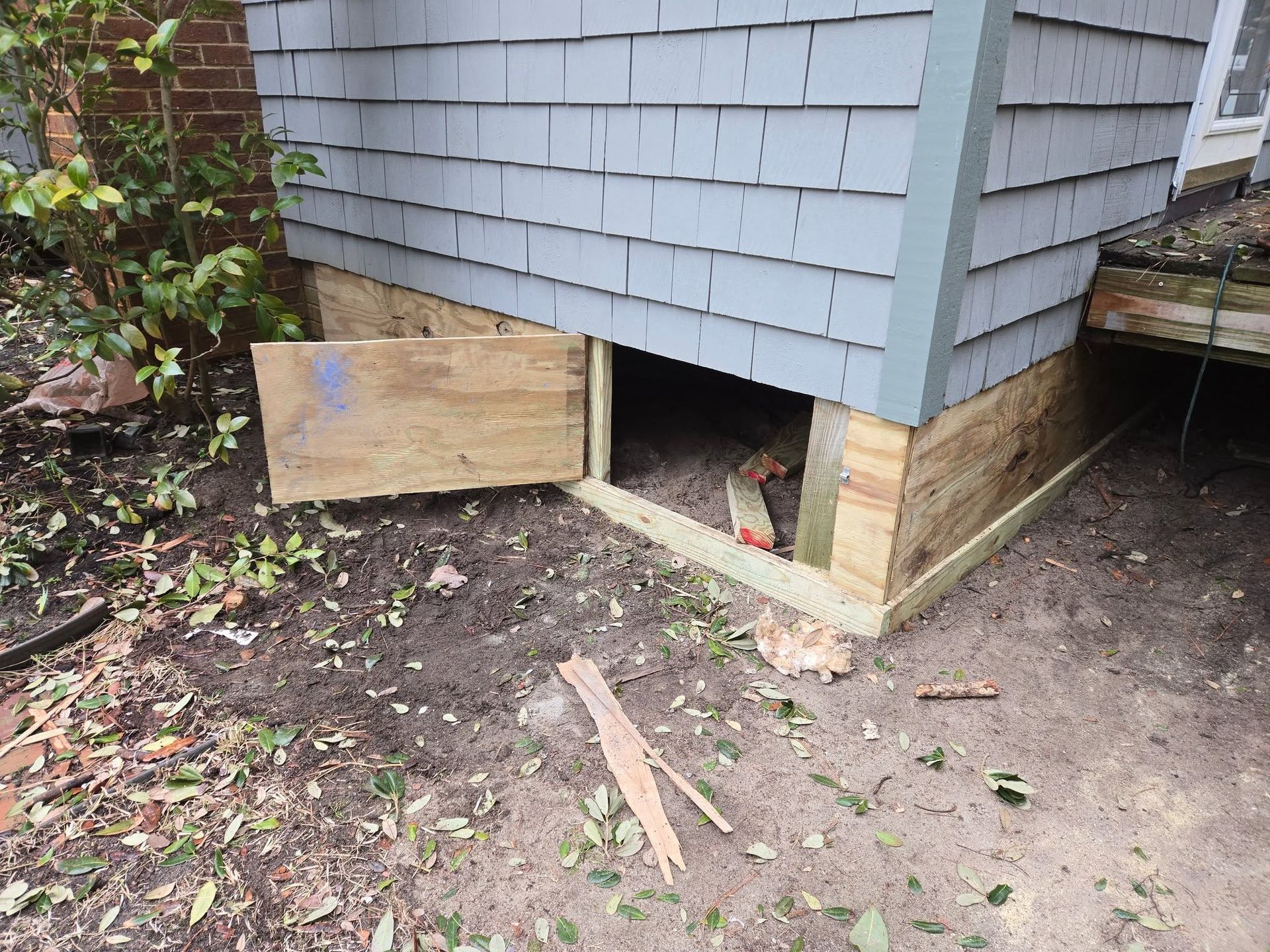 House corner with exposed foundation and a plywood board blocking a crawlspace opening.