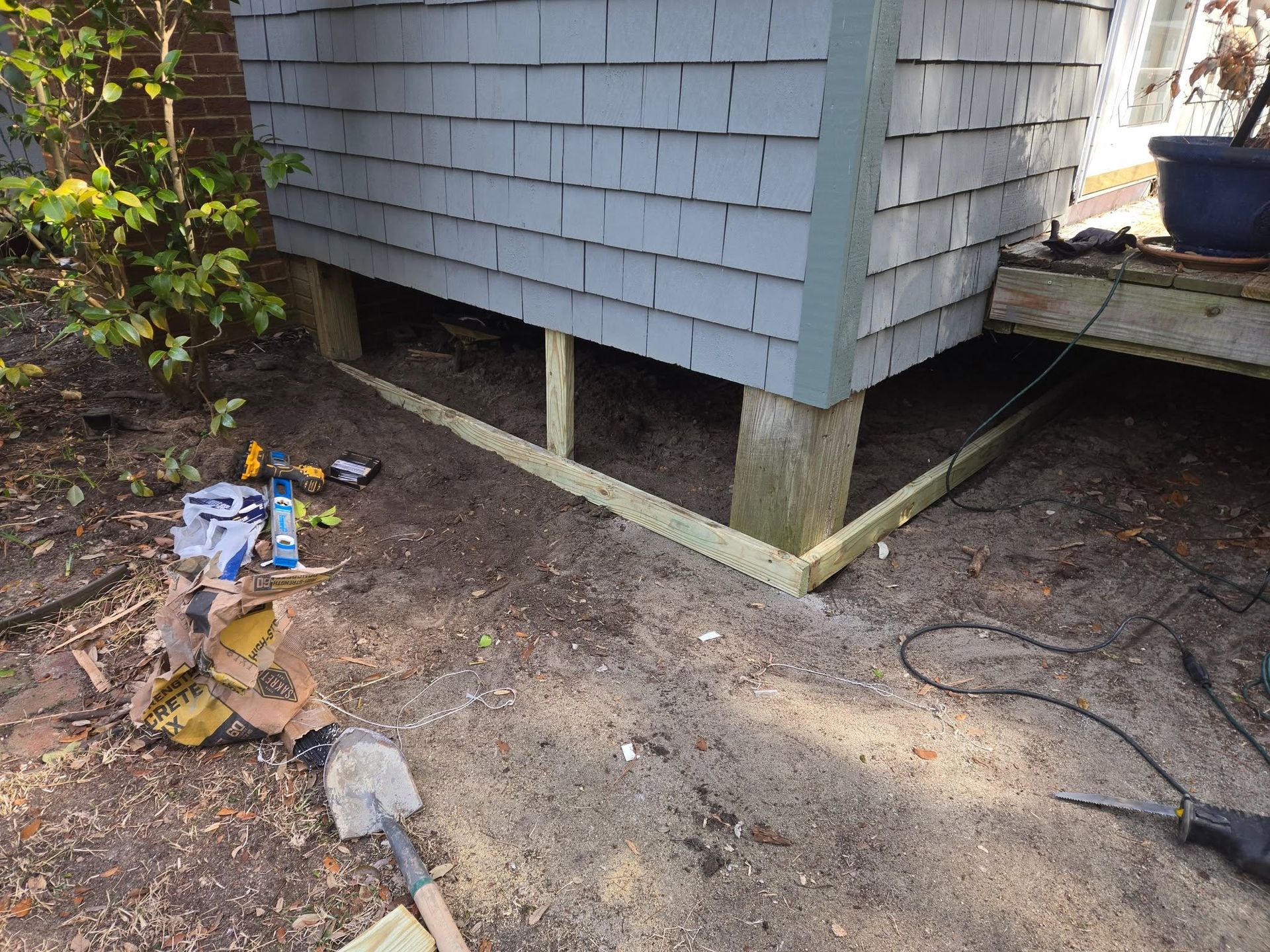 House foundation with exposed supports, dirt ground, and scattered tools beside a deck.