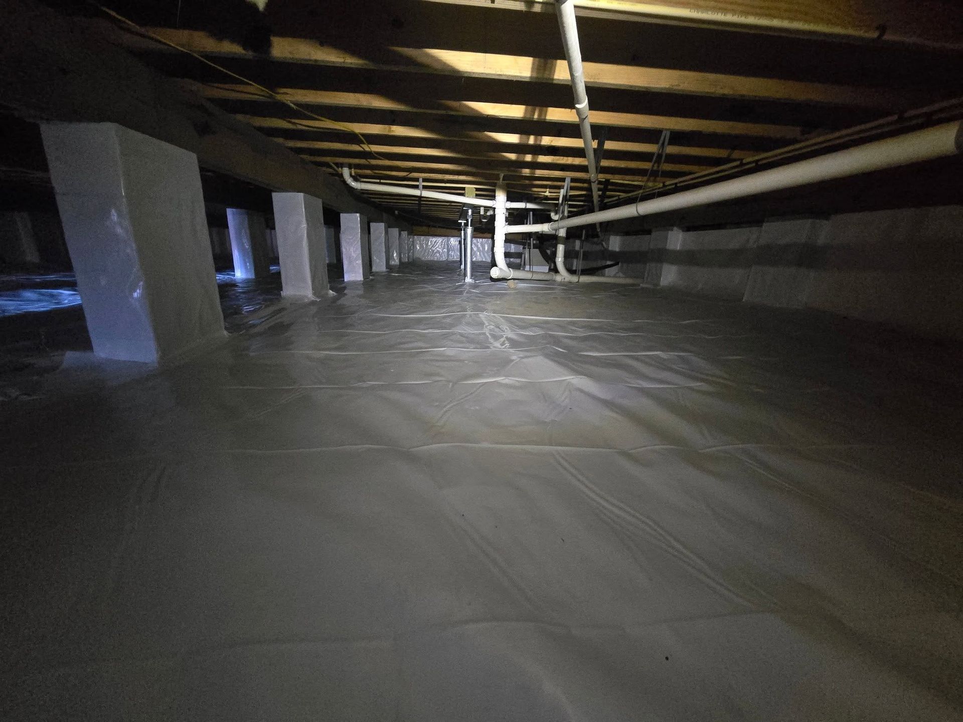 A dim crawlspace with exposed beams, insulation, and a plastic-covered floor illuminated by a flashlight.