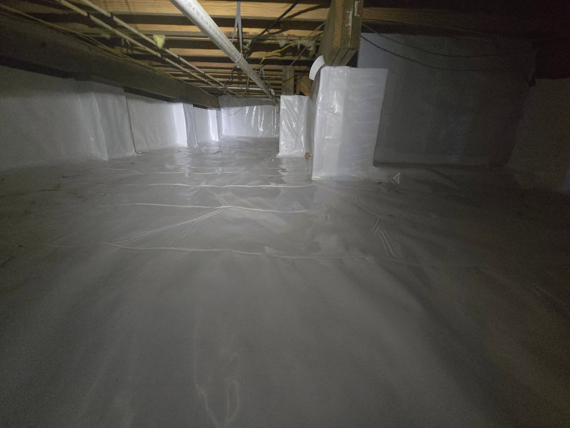 Insulated crawl space with white vapor barrier lining the floor and walls under exposed wooden beams