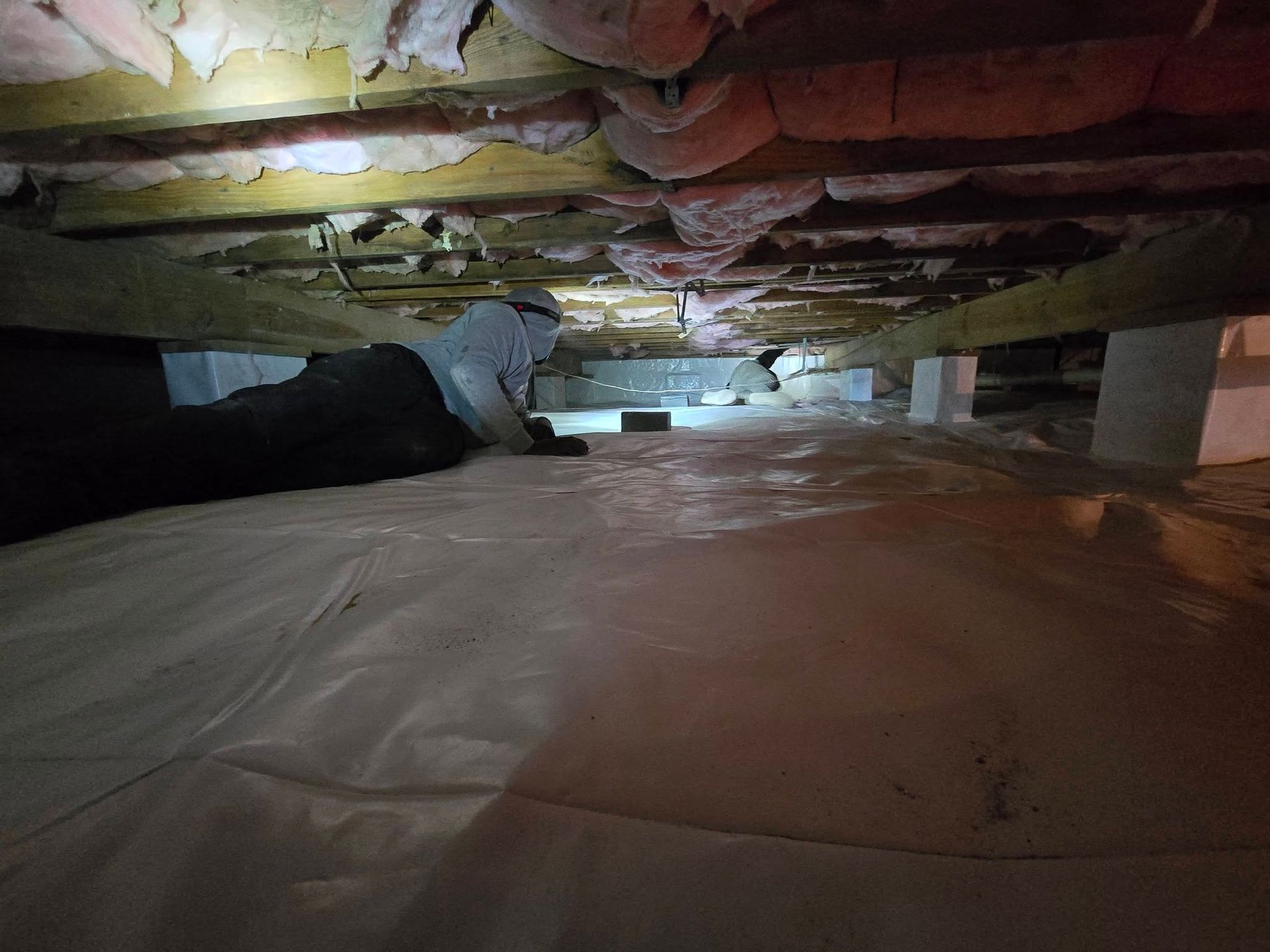 Crawlspace under a house with exposed insulation, ducts, and a plastic vapor barrier on the floor.