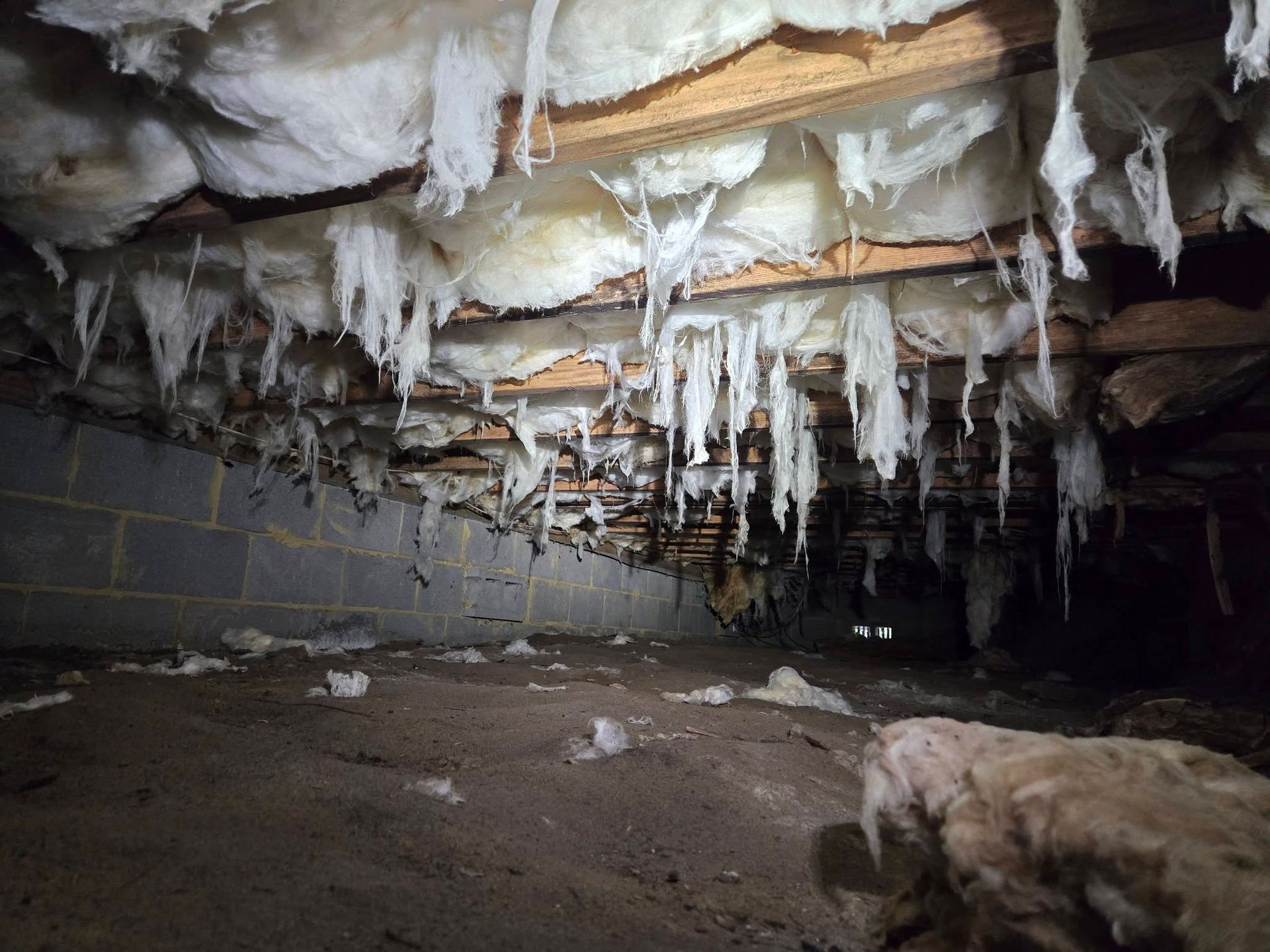 Low crawlspace with hanging insulation and icicles under wooden beams, lit dimly inside a basement.