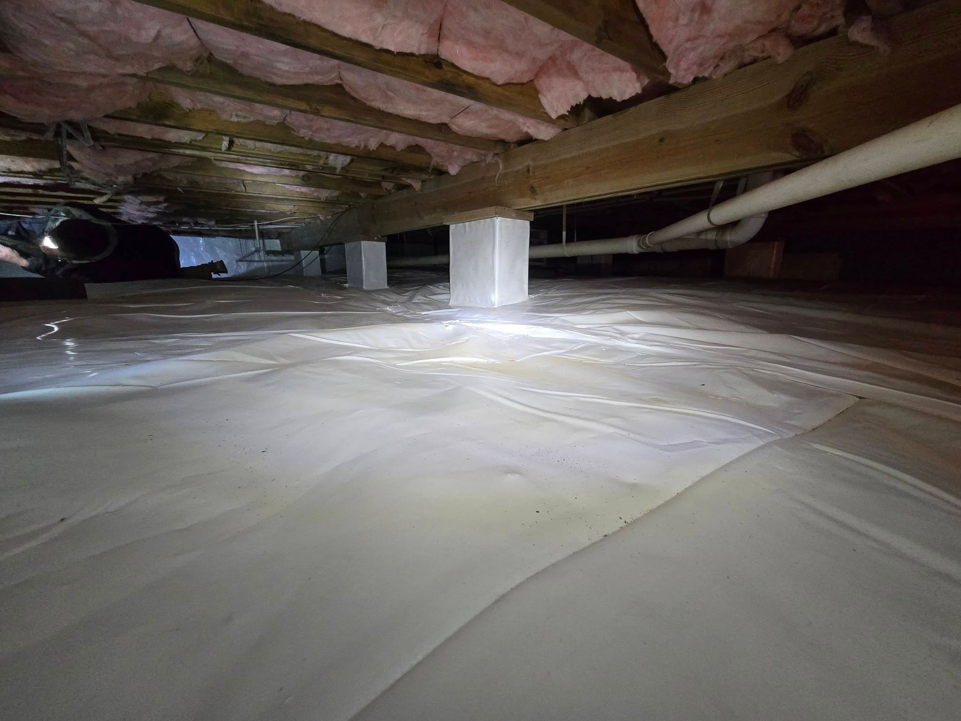 Crawlspace under a house with white plastic vapor barrier, exposed beams, and a support post