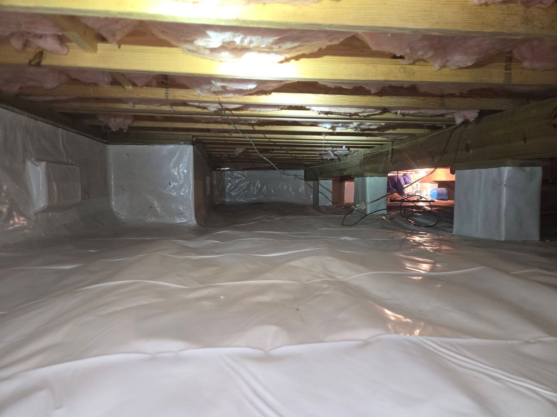 Crawlspace with plastic vapor barrier on floor, wrapped ducts and joists overhead, and a small lit opening ahead