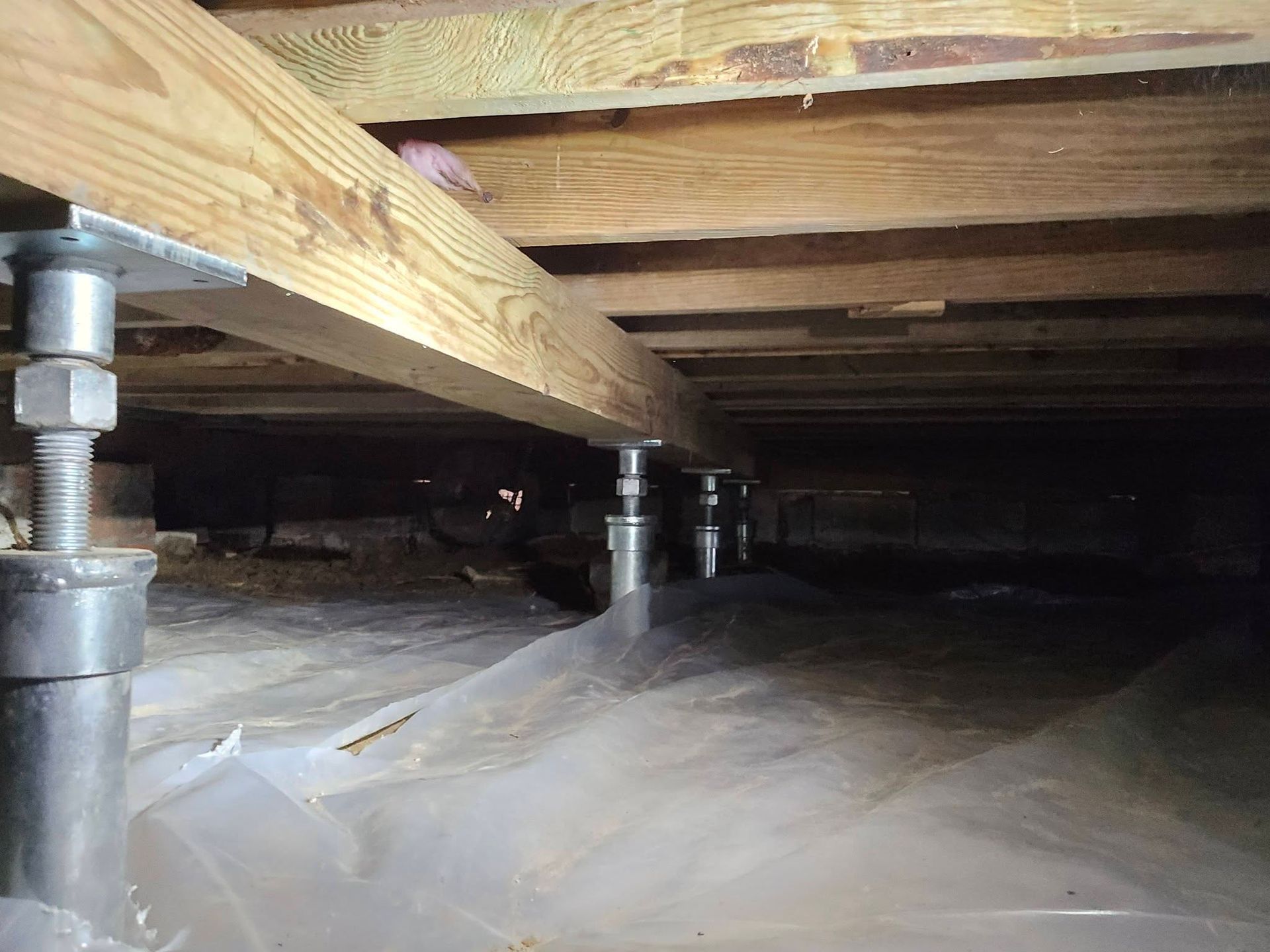 Crawlspace under house with wooden beams, metal supports, and plastic sheeting on the ground