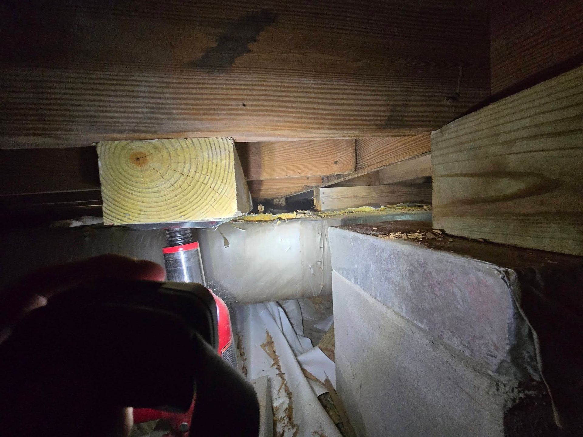 Flashlight inside a crawlspace shows wooden joists, insulation, and a narrow passage with debris.