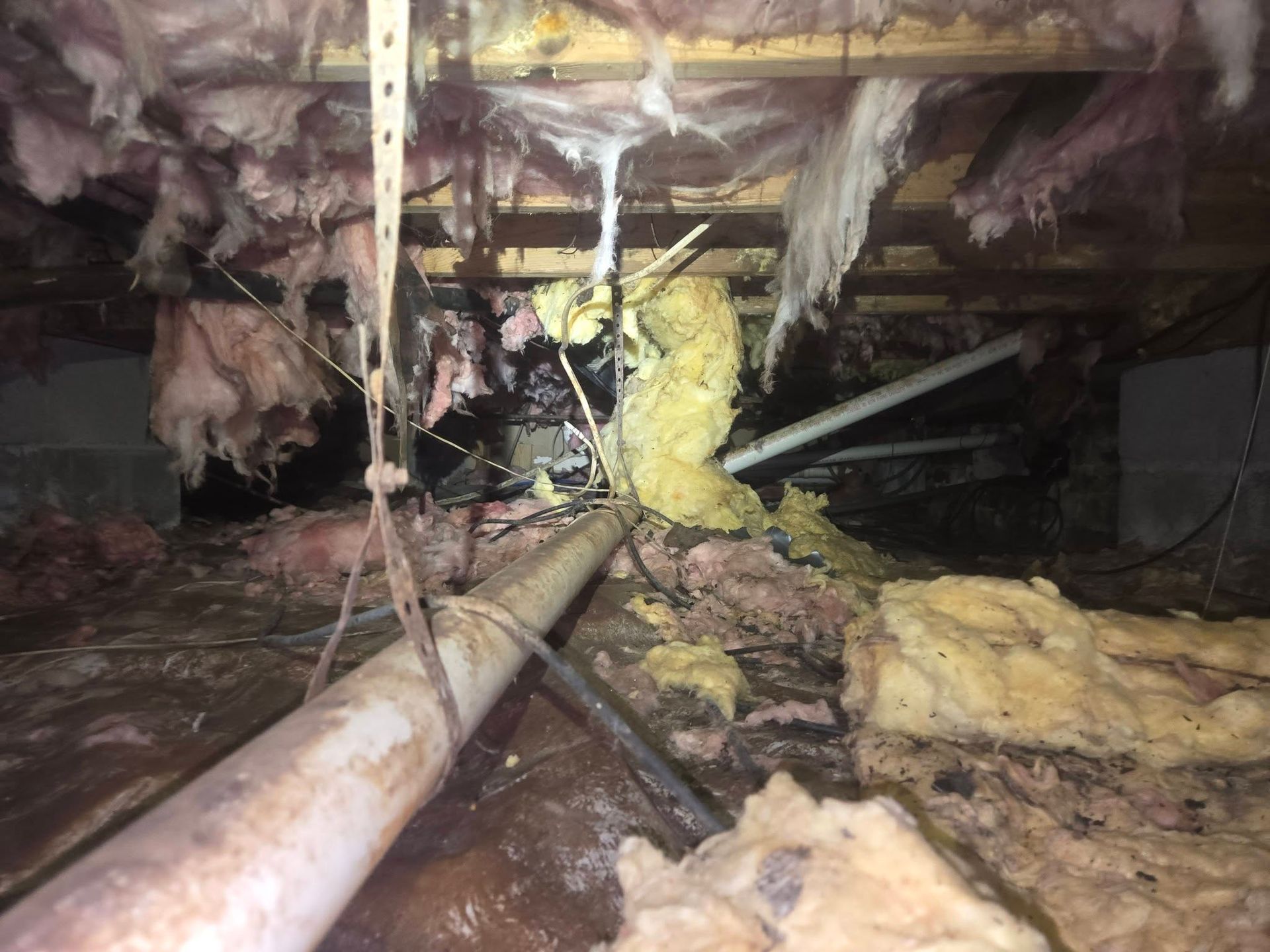 Crawlspace with exposed pipes, insulation, and damaged floor joists beneath a building
