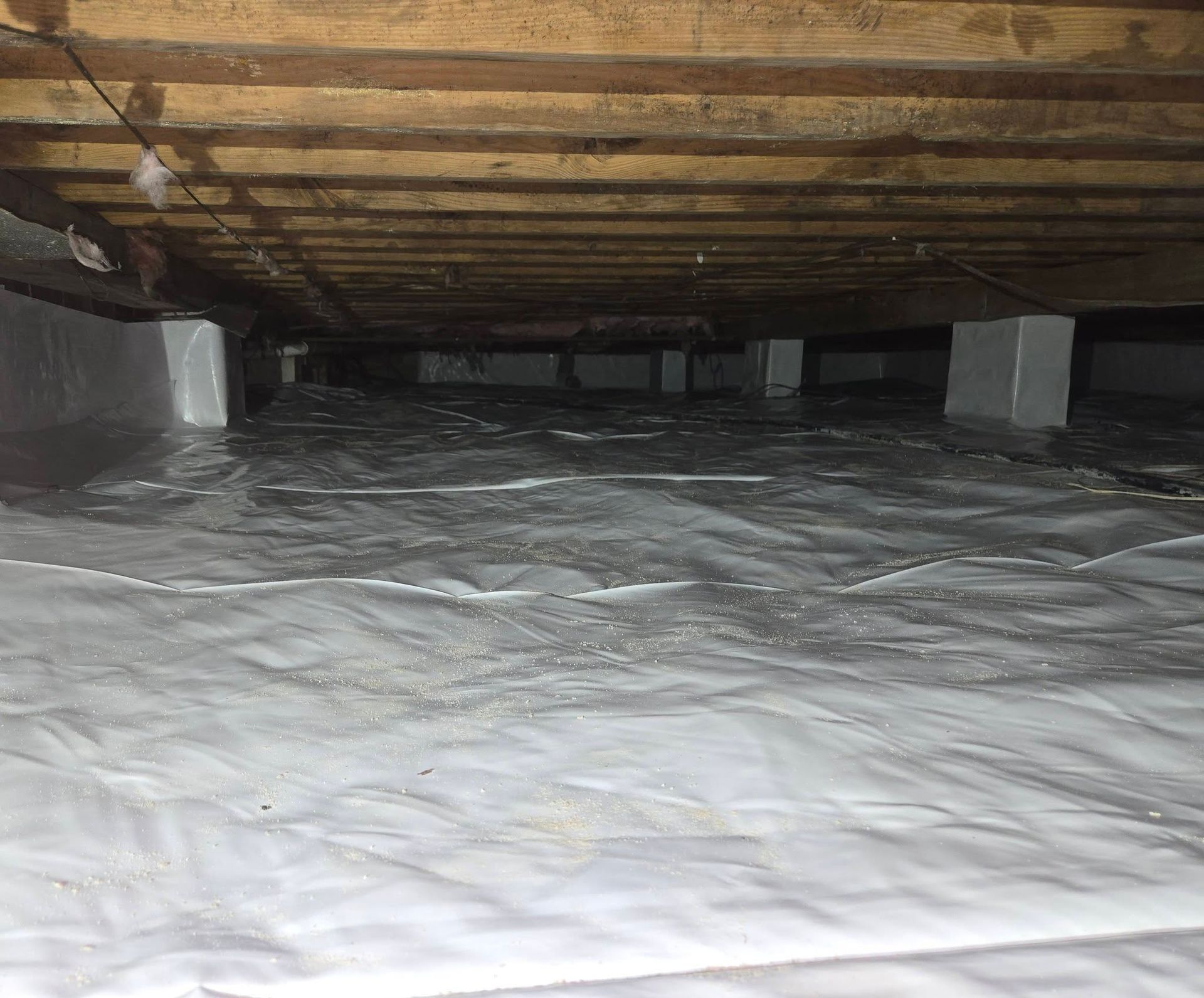Crawlspace under a house with wooden beams, cinder block supports, and white plastic vapor barrier on the ground