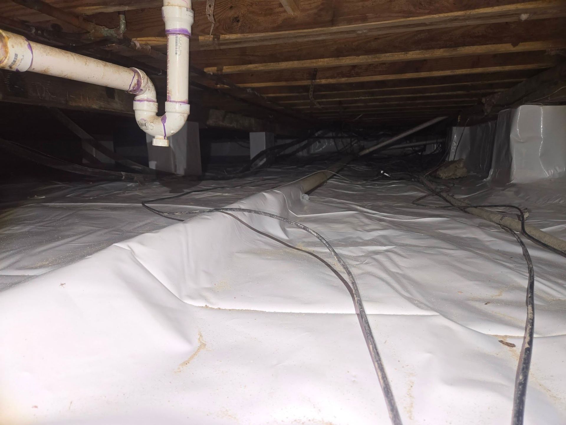 Crawlspace under a house with plastic vapor barrier, exposed pipes, and wooden joists.