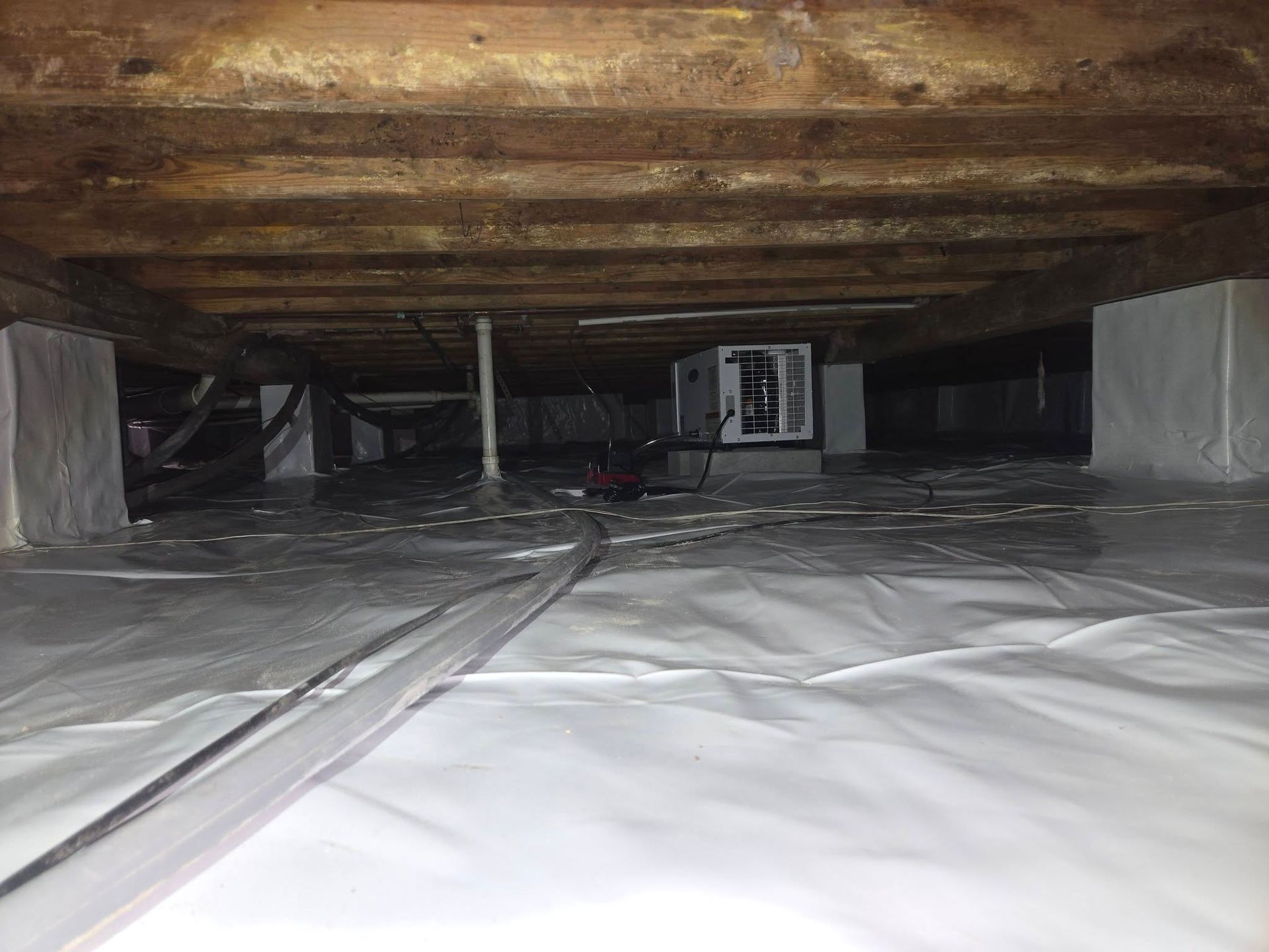Low crawlspace with white vapor barrier, exposed wooden joists, and a small HVAC unit in the back