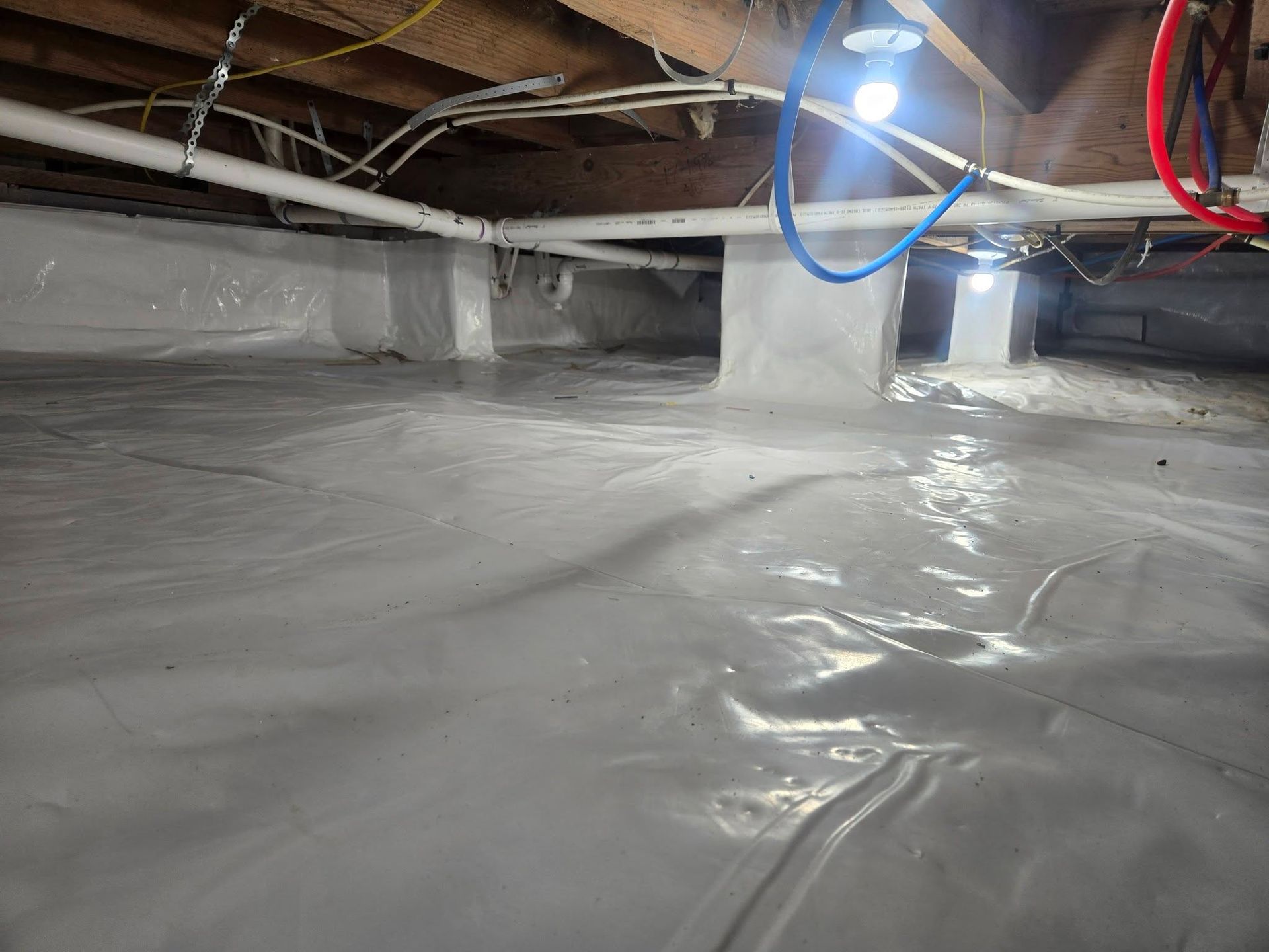 Plastic-covered crawl space with exposed beams and pipes, lit by a bright work light