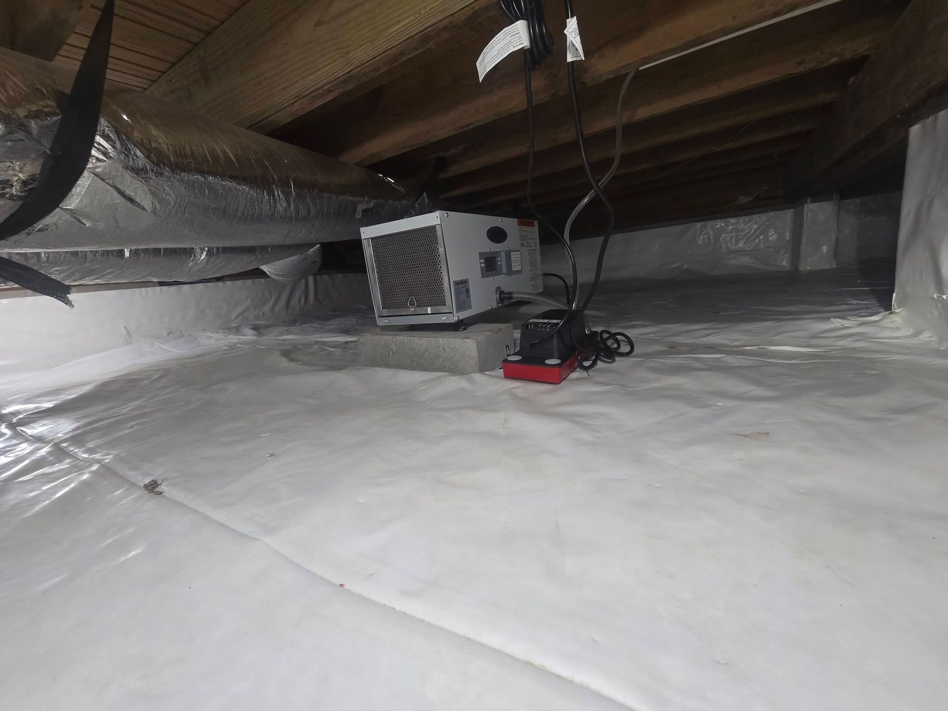 Attic with white insulation, exposed beams, and a gray HVAC unit on the floor under the roof.