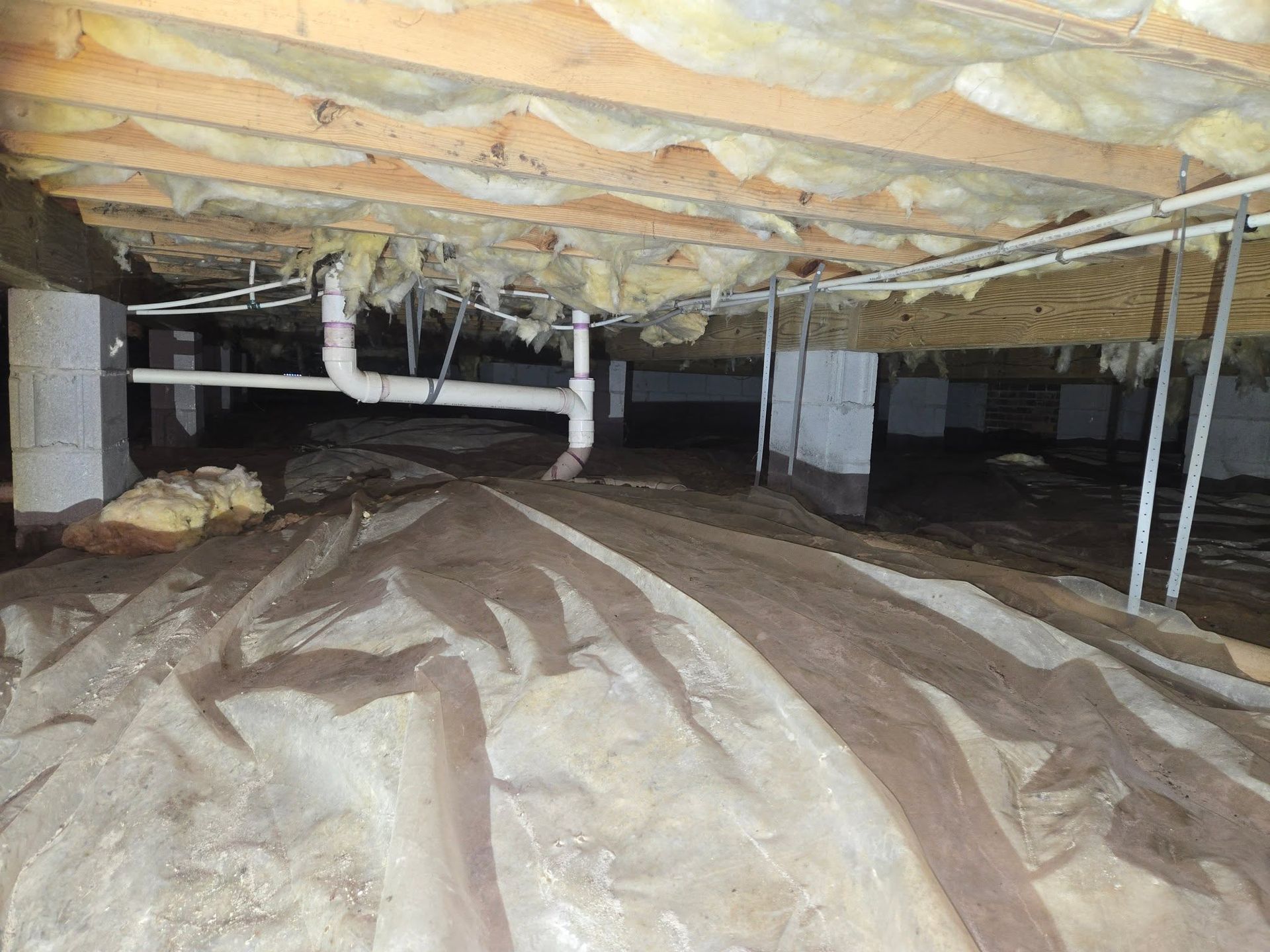 Crawlspace under a house with exposed insulation, pipes, and plastic vapor barrier on the floor