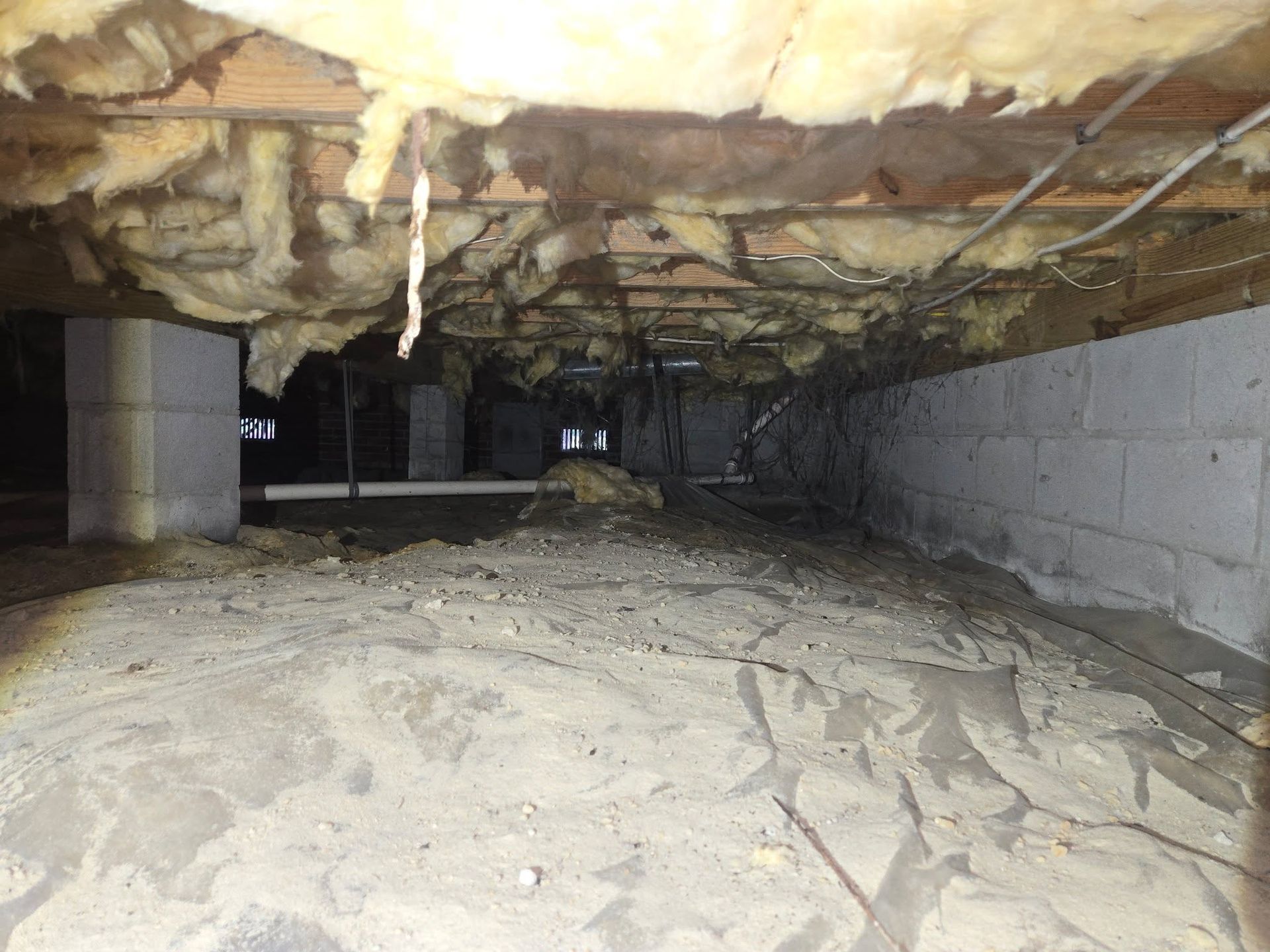 Low crawlspace with insulation, exposed pipes, and a dirt floor under a house