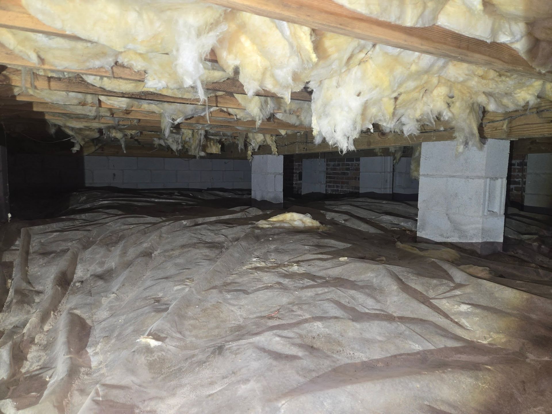 Crawlspace with exposed insulation, support beams, and plastic-covered ground under a house
