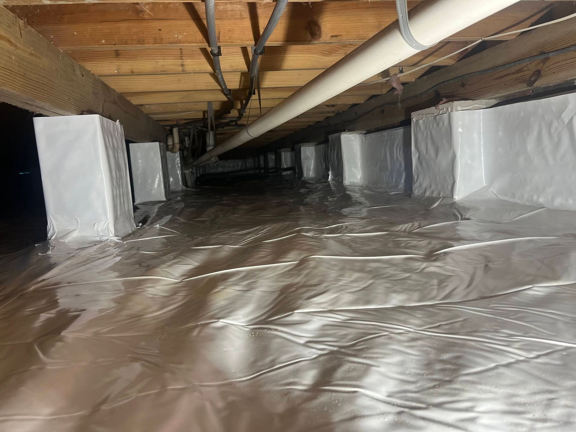 Crawlspace with insulated walls, exposed beams, and plastic vapor barrier on the ground.