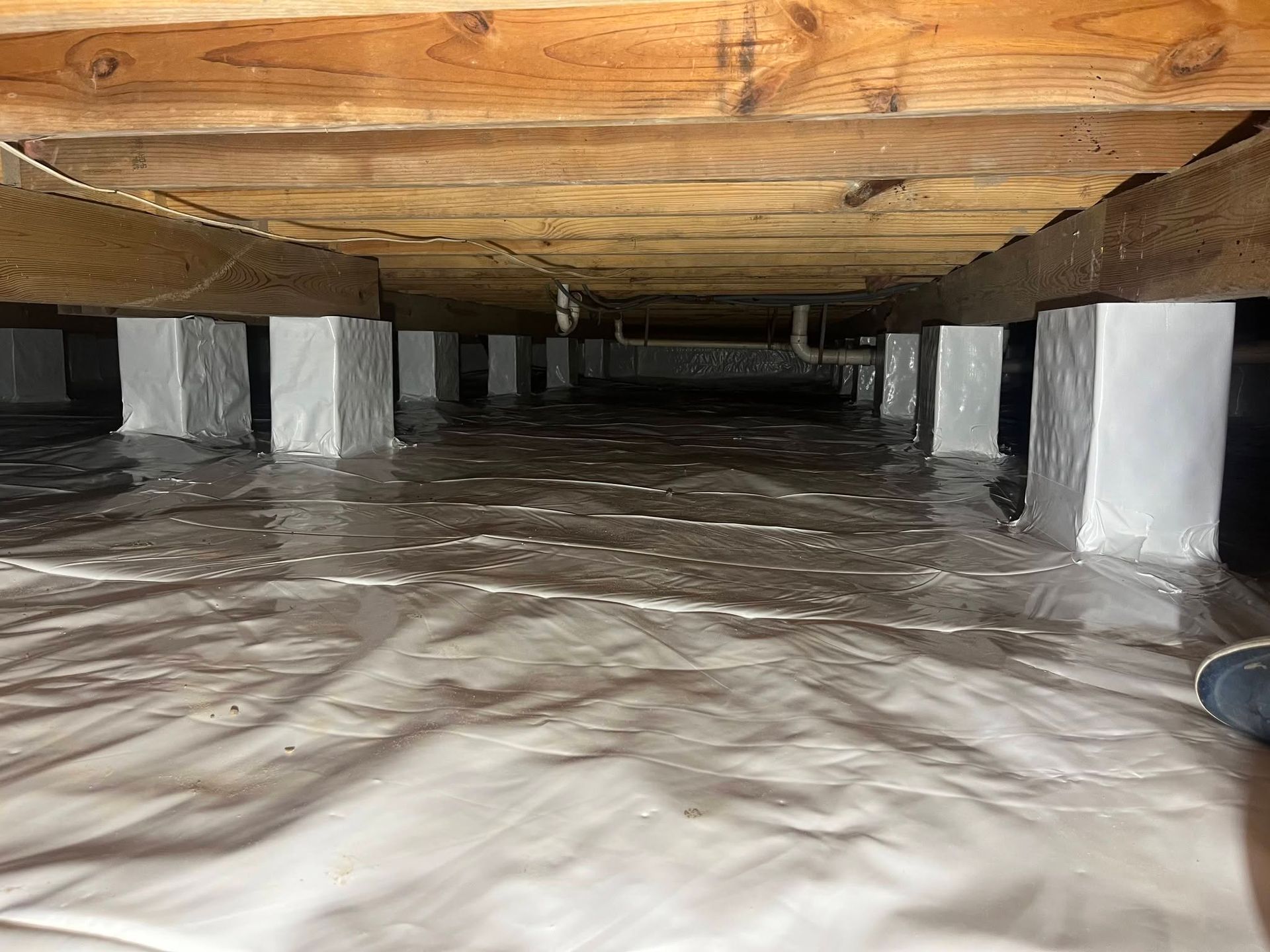 Low crawlspace under a wooden floor with white support blocks and plastic sheeting on the ground