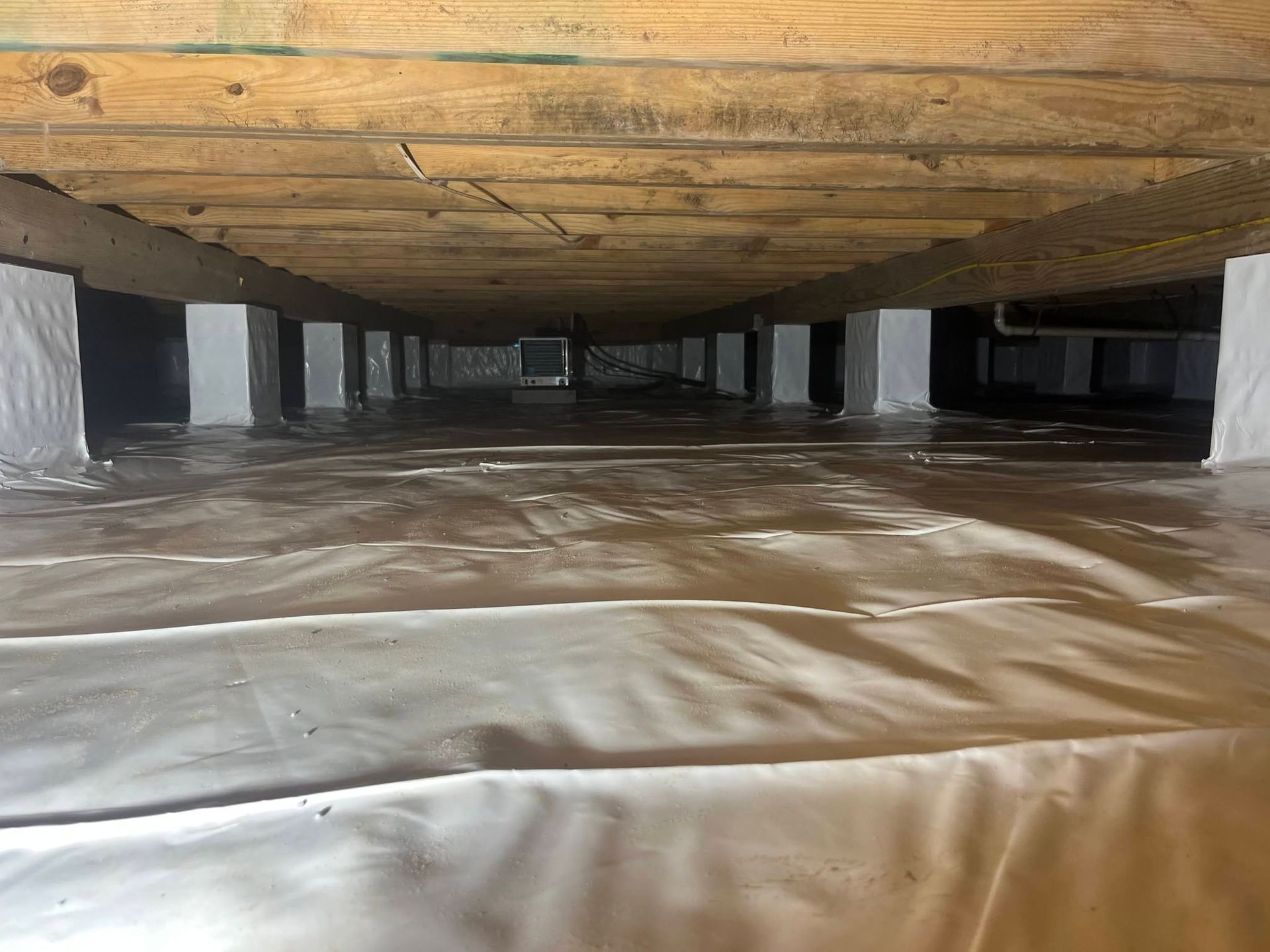 Crawlspace under a wooden floor with insulation and support posts, viewed from a low angle