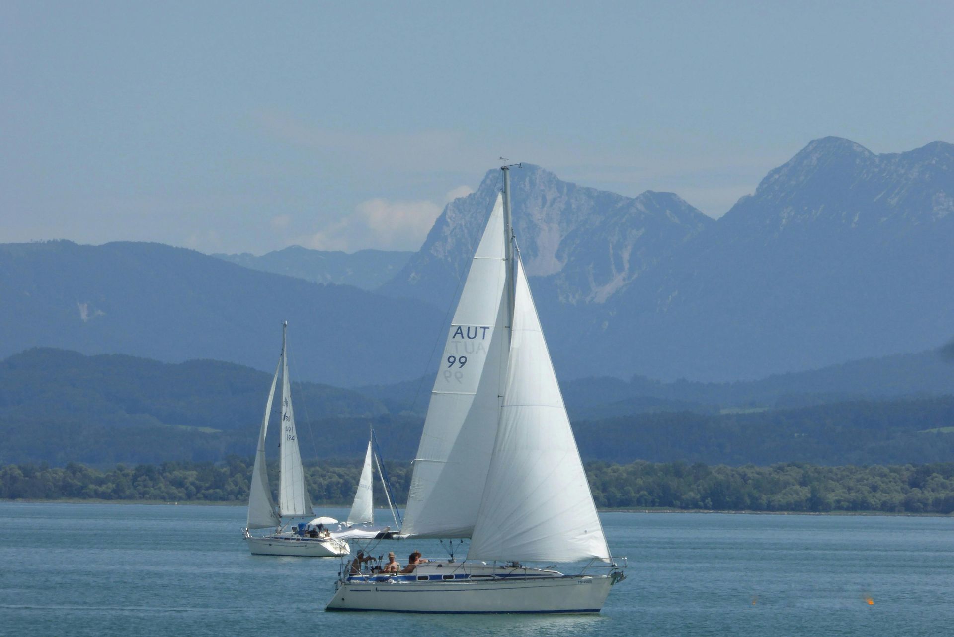 Ein Segelboot mit weißen Segeln gleitet auf ruhigem, blauem Wasser; im Hintergrund sind Berge zu sehen.