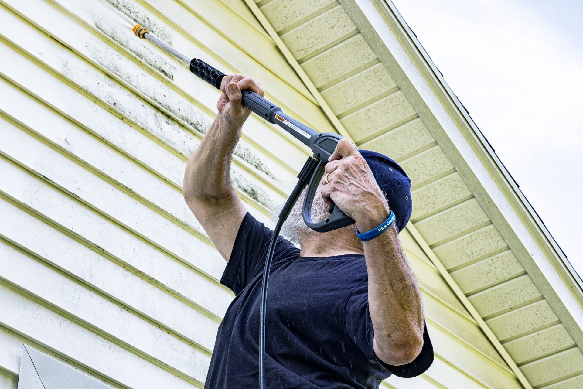 Senior man using residential pressure washing service to clean home siding with high-pressure water.