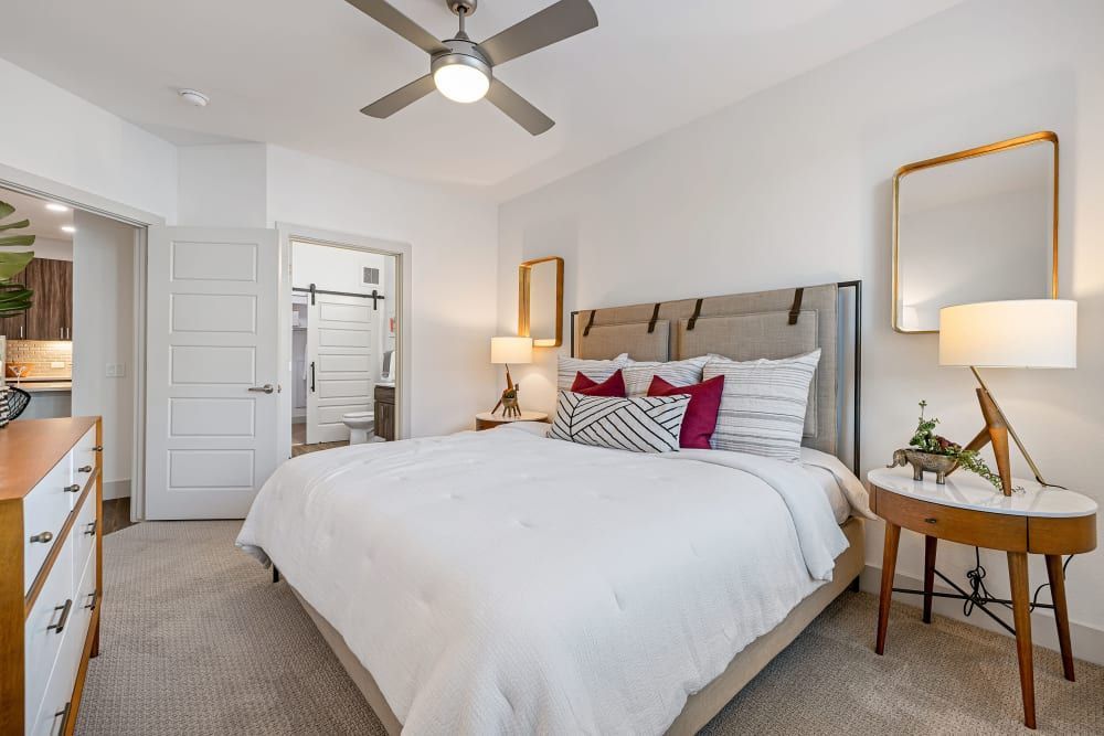 Bright modern bedroom with white bed, wooden nightstands, ceiling fan, and a door to the hallway.
