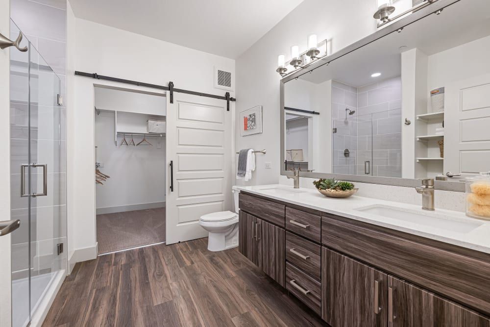 Bright modern bathroom with double vanity, white tile, large mirror, and walk-in closet visible through open door.
