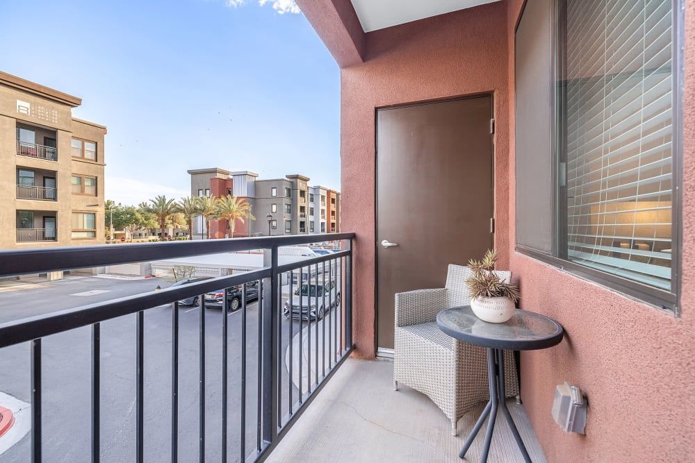 Small apartment balcony with railing, door, window blinds, and a side table with decor overlooking nearby buildings