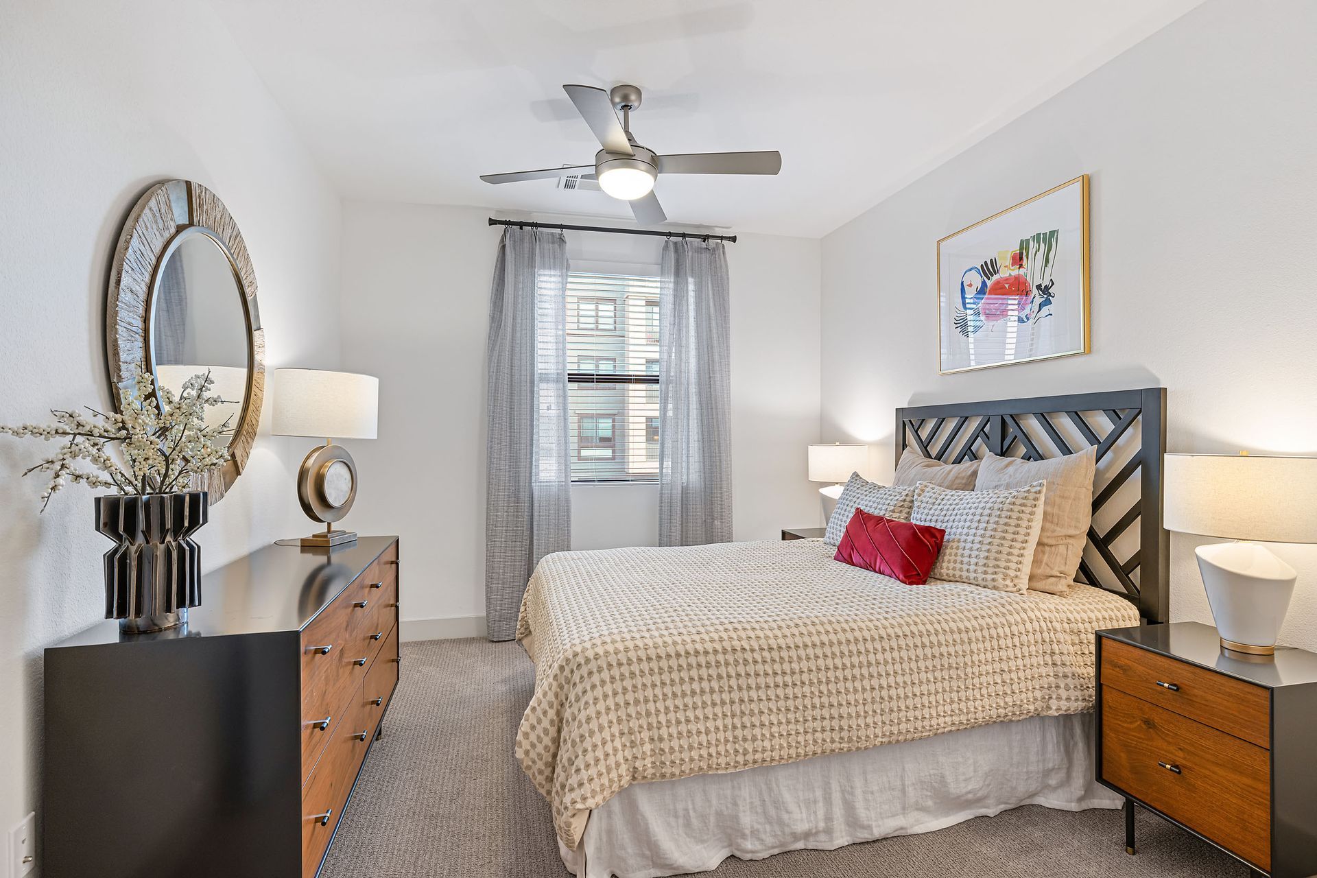 Bright bedroom with a gray bed, wooden dressers, lamps, and a ceiling fan under a window