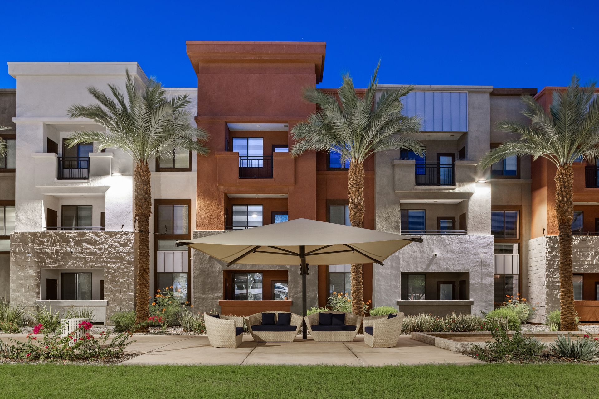 Modern apartment building with stone and wood facade, palm trees, and patio umbrellas in front