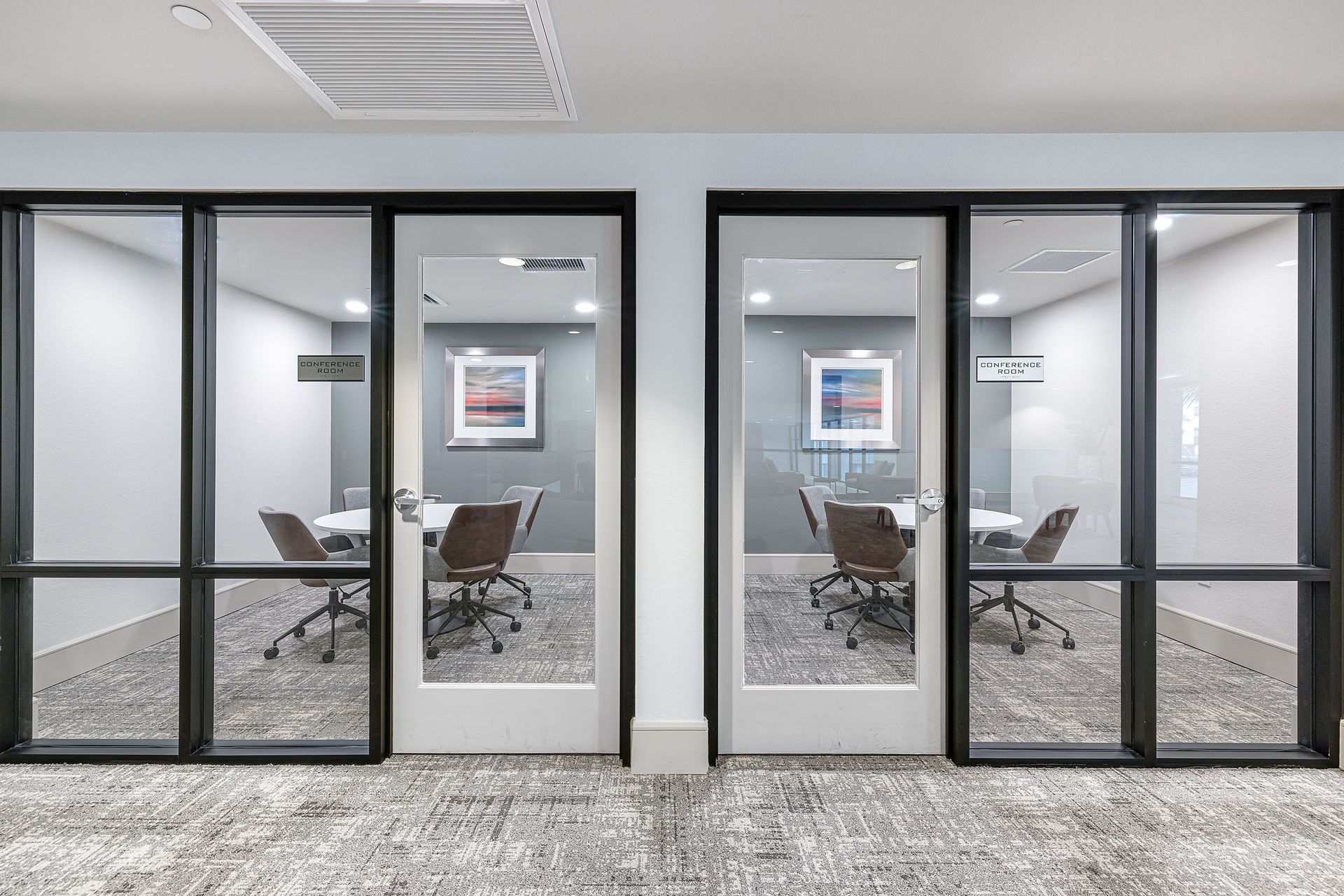 Modern office conference room with glass walls, chairs, framed wall art, and gray carpet