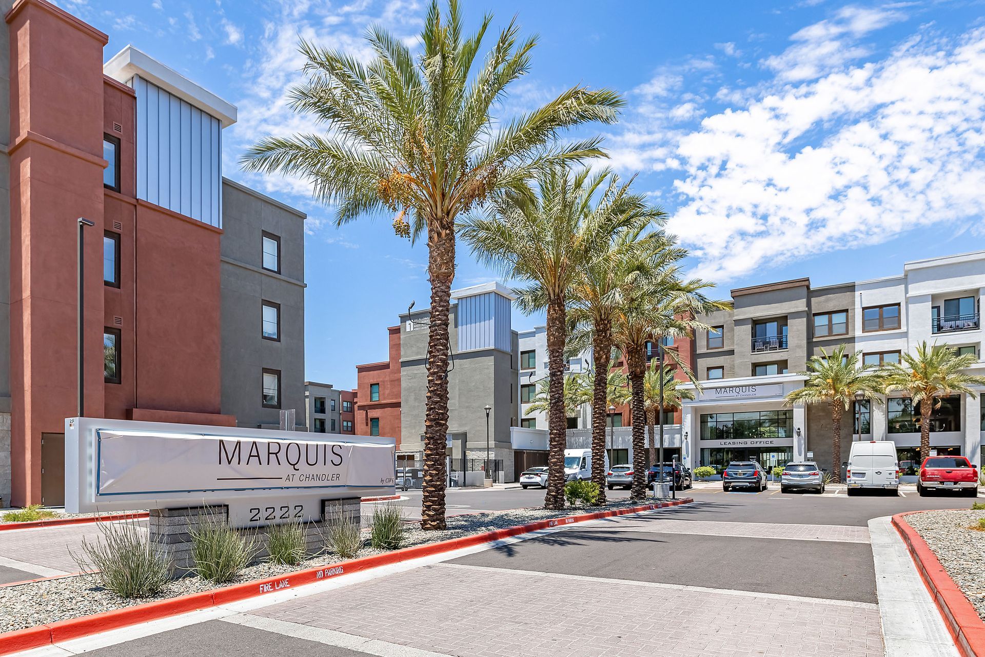Modern apartment complex entrance with palm trees, parking area, and brick-and-gray buildings under a blue sky