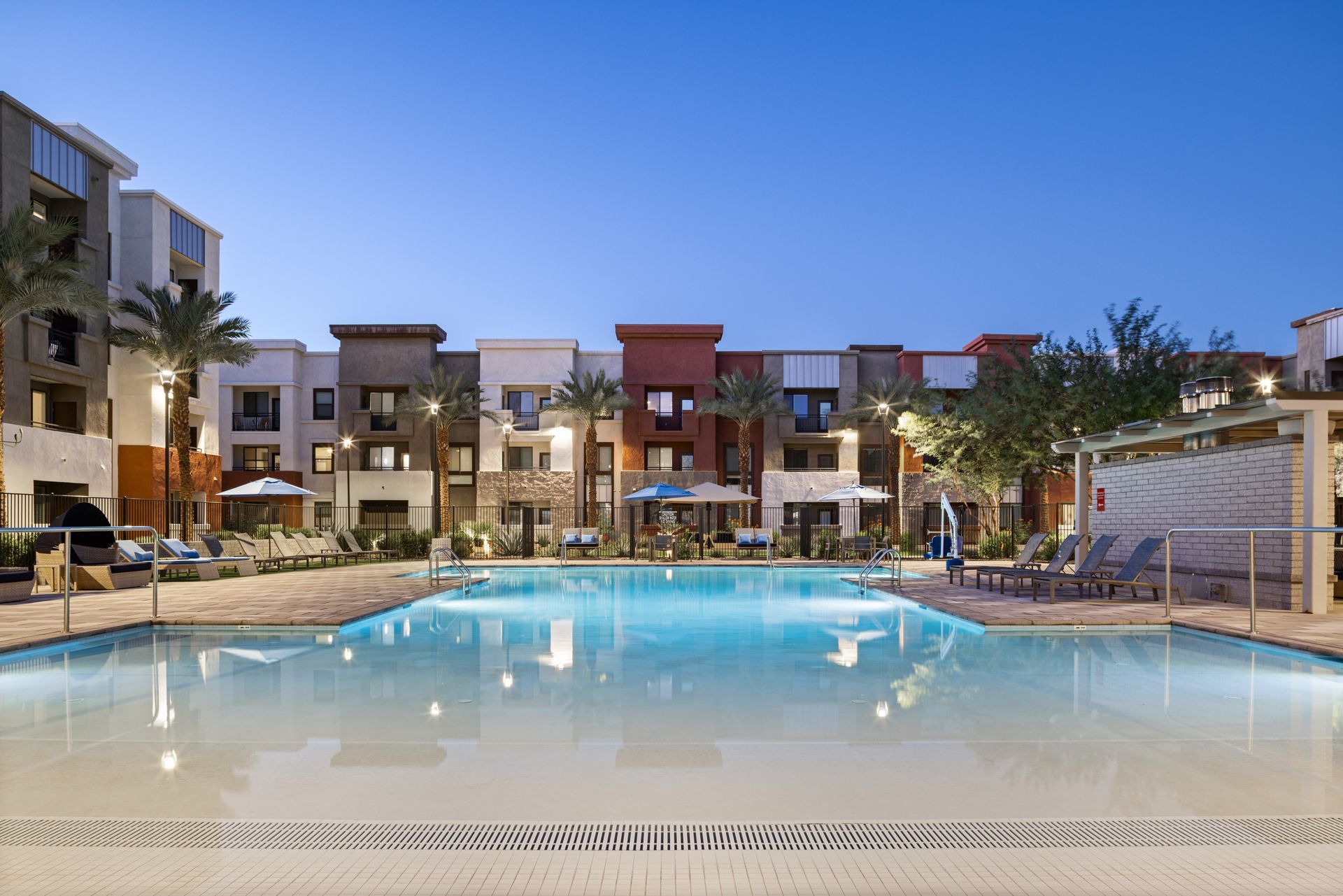 Modern apartment complex pool at dusk with lit buildings and lounge chairs
