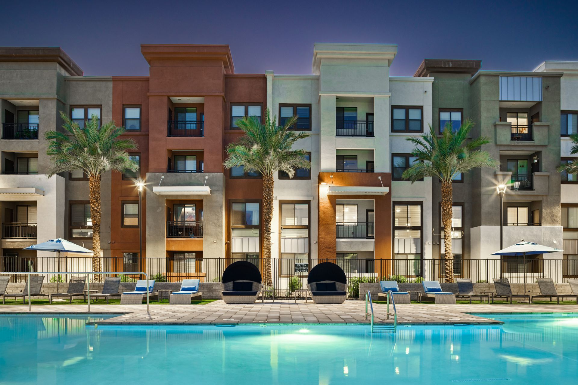 Modern apartment complex with palm trees and a turquoise pool at dusk