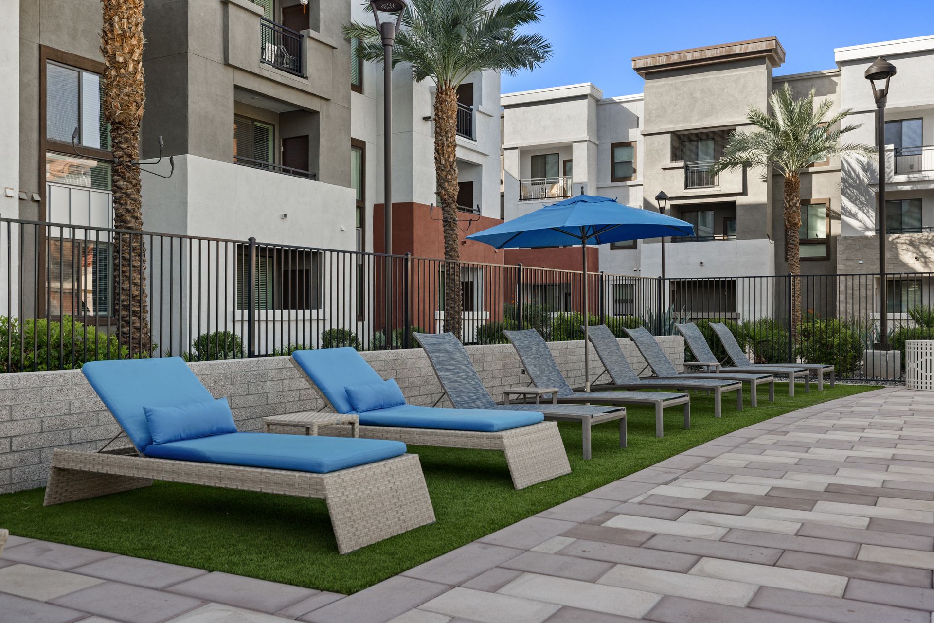 Apartment courtyard with blue lounge chairs, umbrella, and paved walkway beside buildings