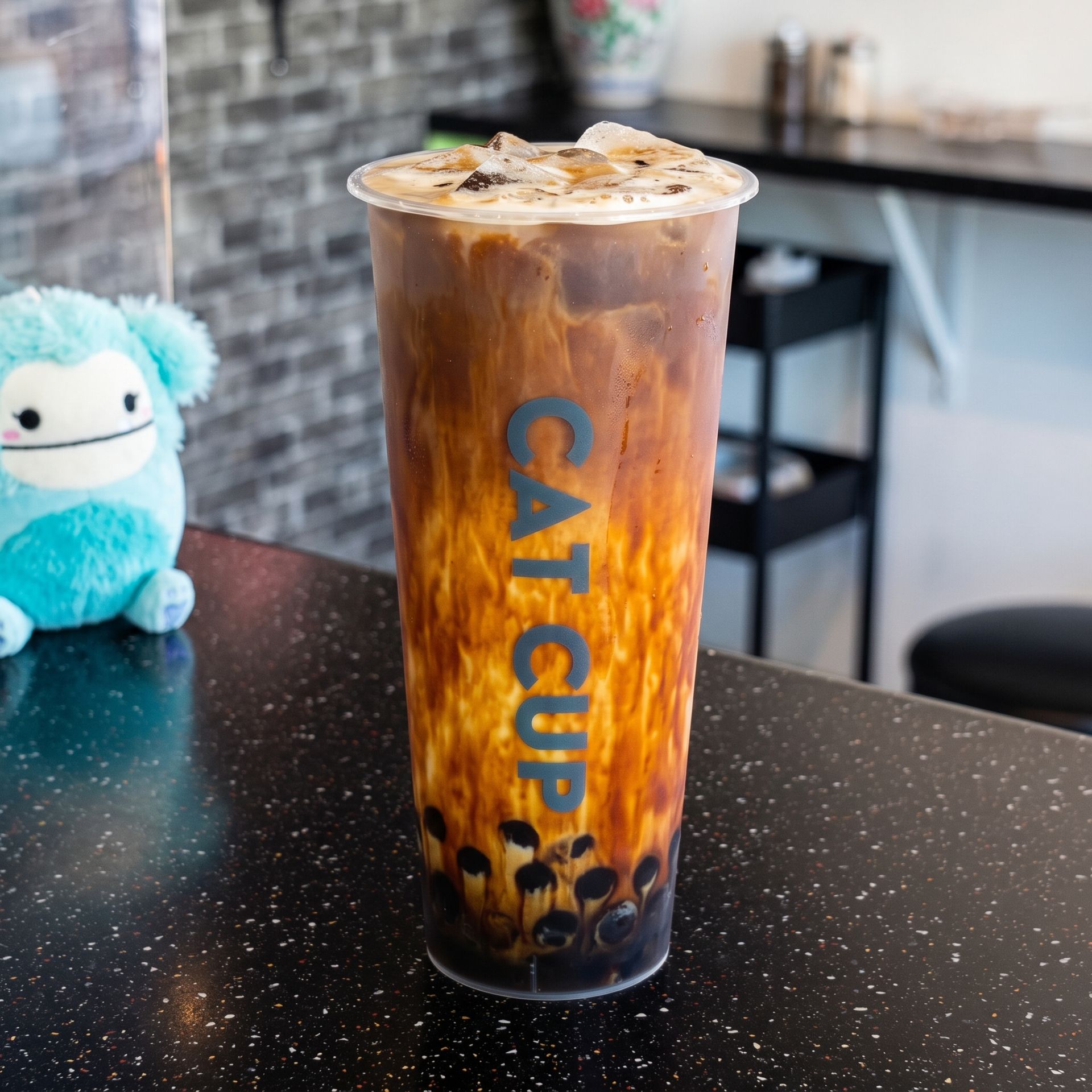 Iced bubble tea in a clear cup with blue “CAT CUP” text on a black table, beside a small blue plush toy