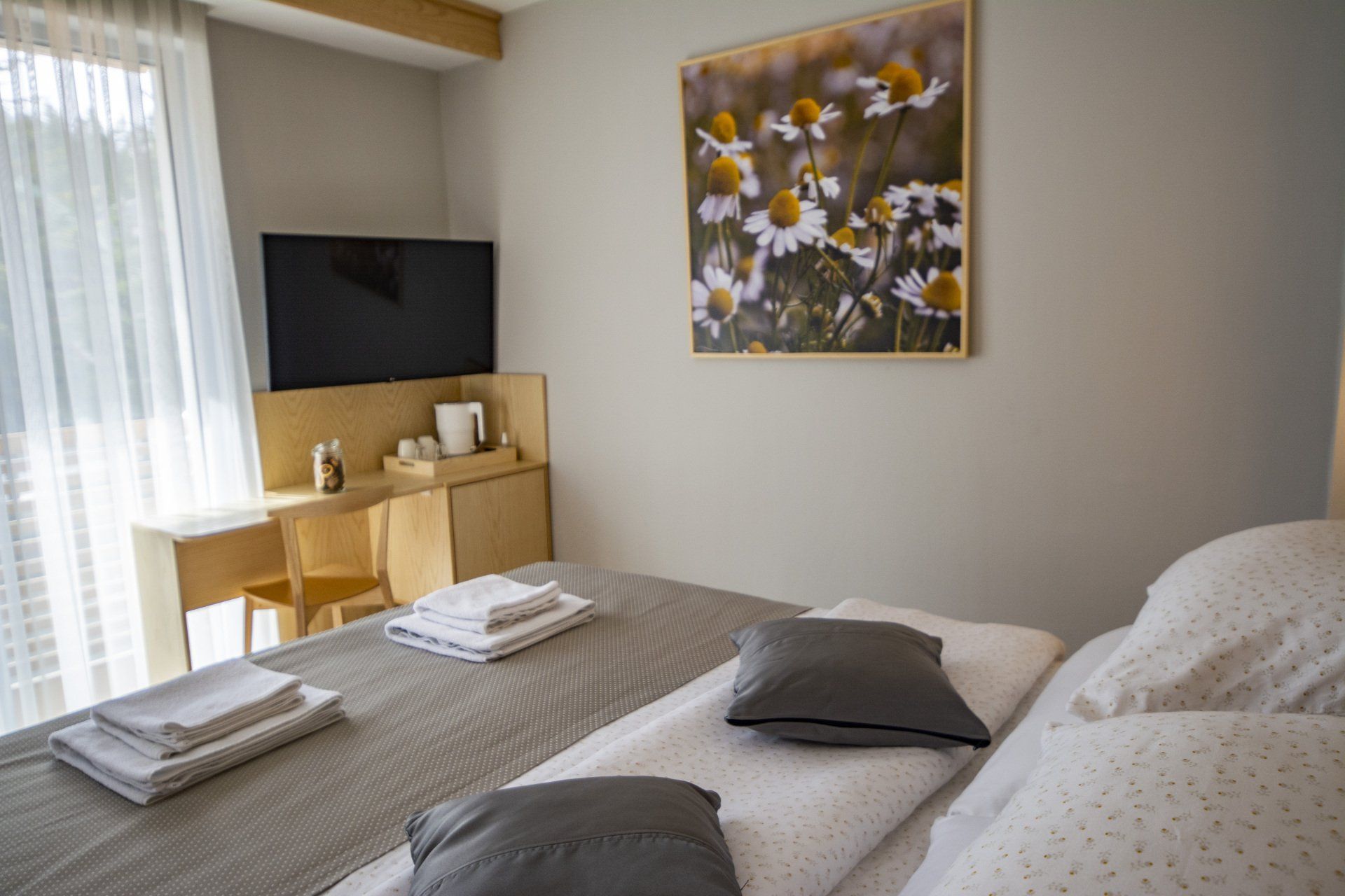 A hotel room with a bed, desk, television and a picture of daisies on the wall.