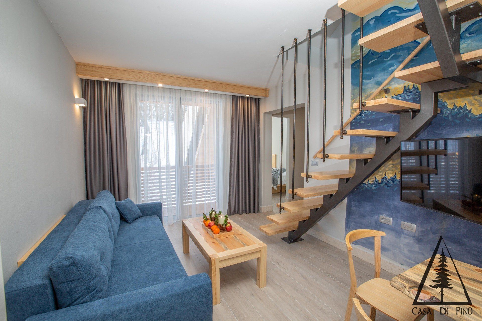 A living room with a blue couch, wooden table and chairs, and stairs and a blue and yellow mural on the wall.