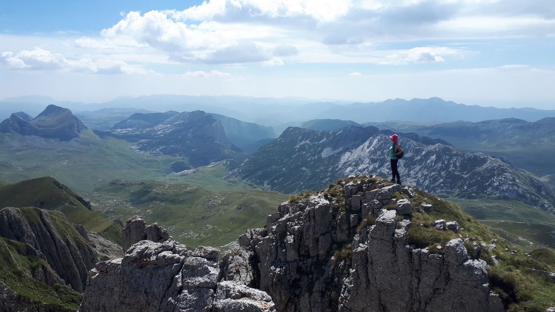 Person stands atop a rocky mountain peak, looking out over a vast, mountainous landscape under a cloudy sky.