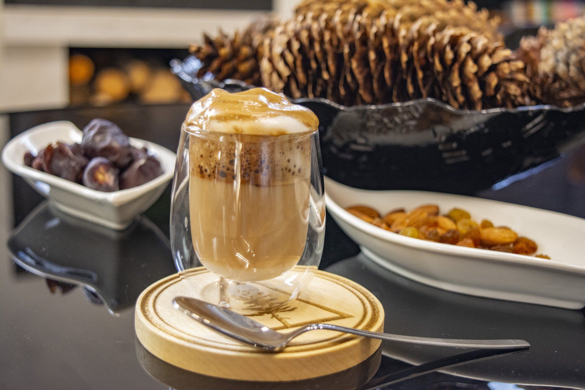 A glass of coffee with a spoon on a wooden coaster on a table.