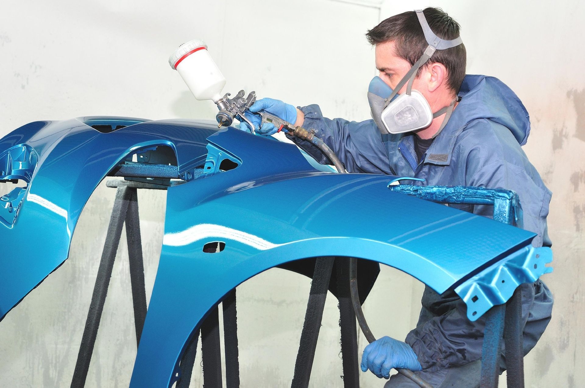 Person in blue overalls spraying blue car parts with paint.
