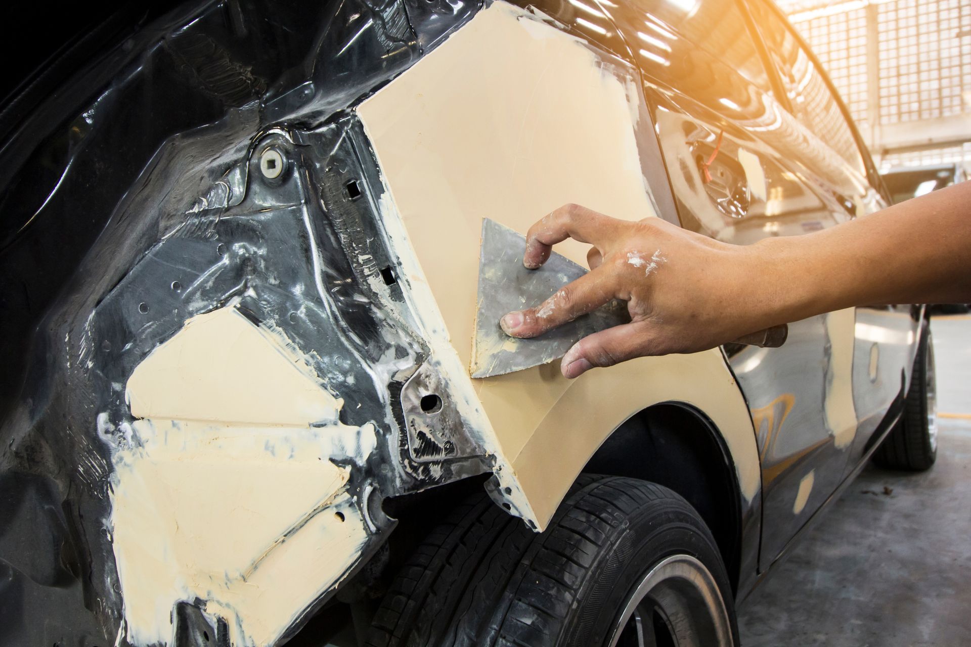 A person sanding body filler on a damaged car panel, in a repair shop. A person sanding body filler on a damaged car panel, in a repair shop.