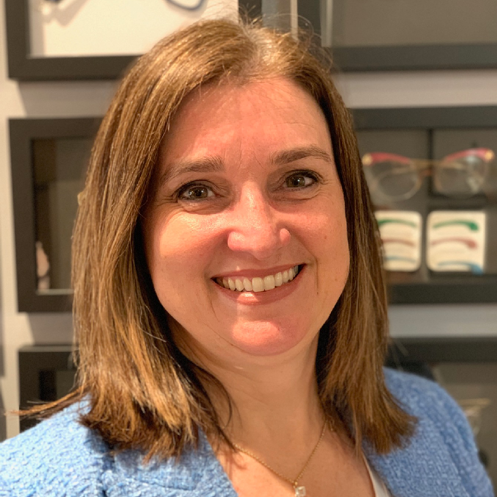 Smiling woman with shoulder-length brown hair in a blue sweater, standing indoors in front of shelves.